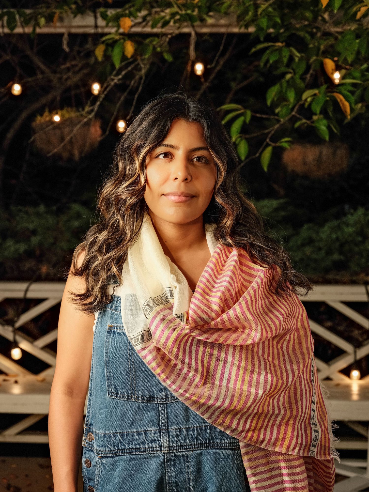Woman in denim overalls with pink striped scarf stands outdoors at night under string lights.