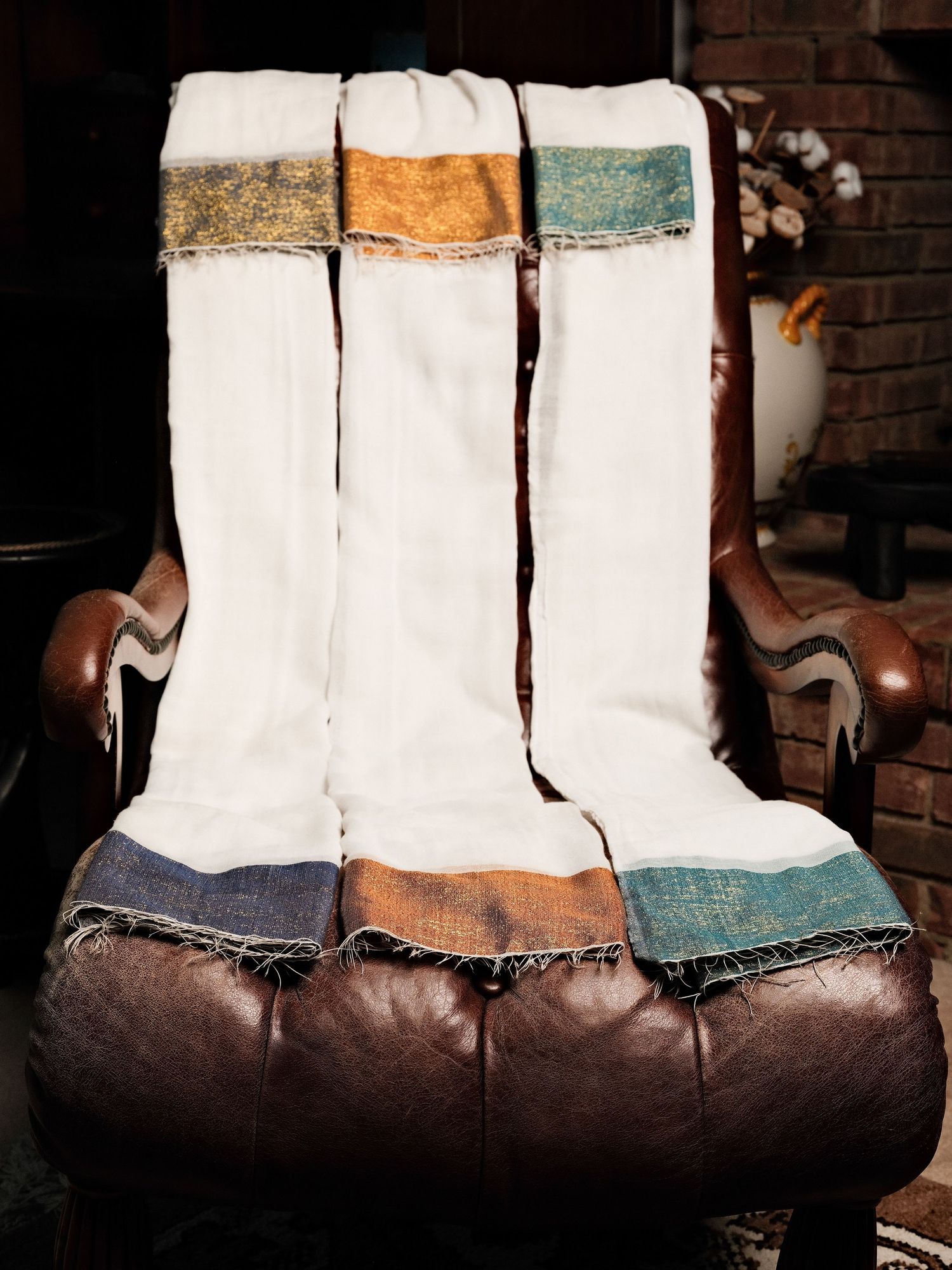 Three white cotton throw blankets with colorful striped ends draped over a brown leather armchair near a fireplace.