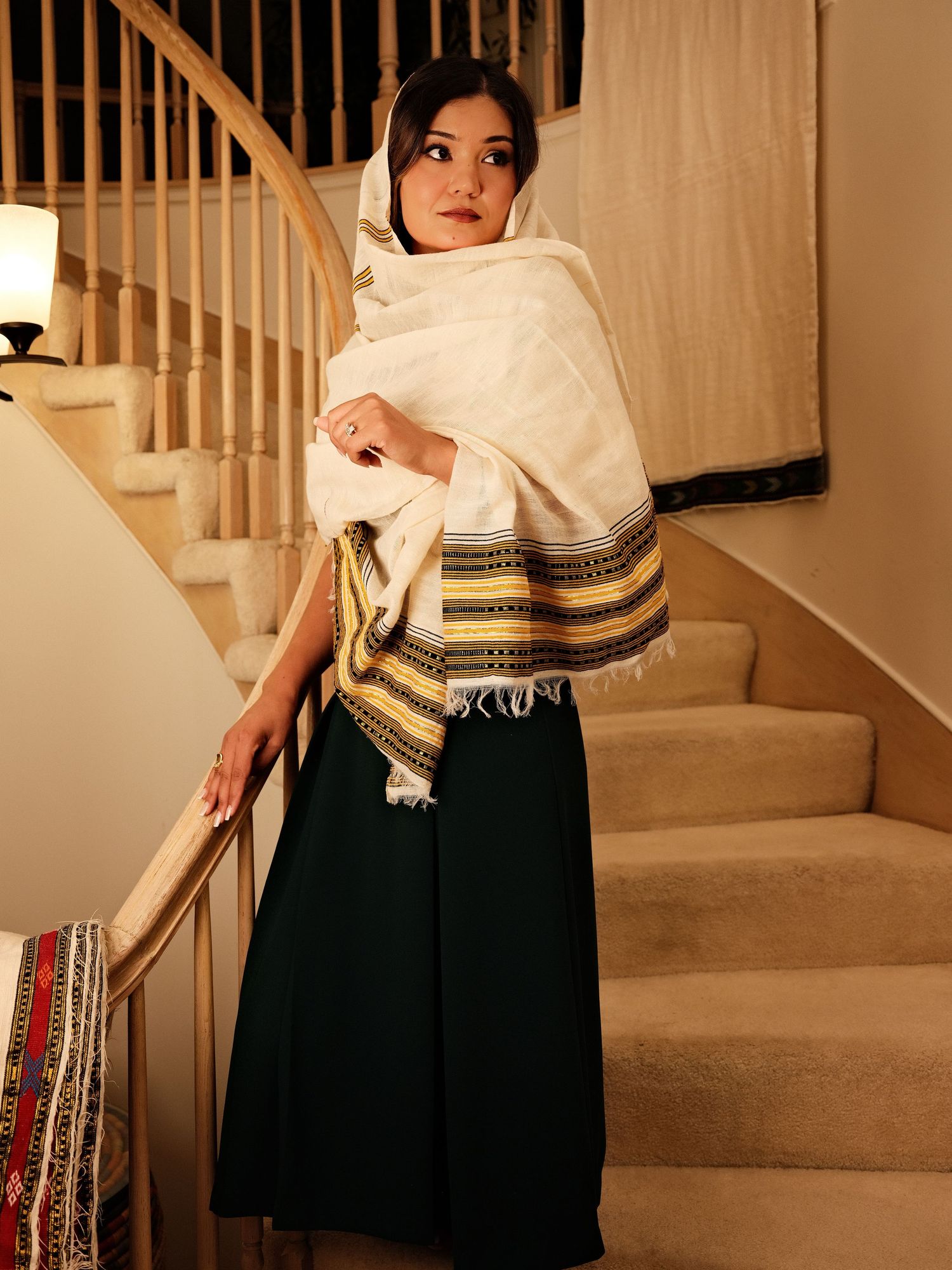 Woman wearing white shawl with gold stripes and dark green maxi skirt standing on staircase.