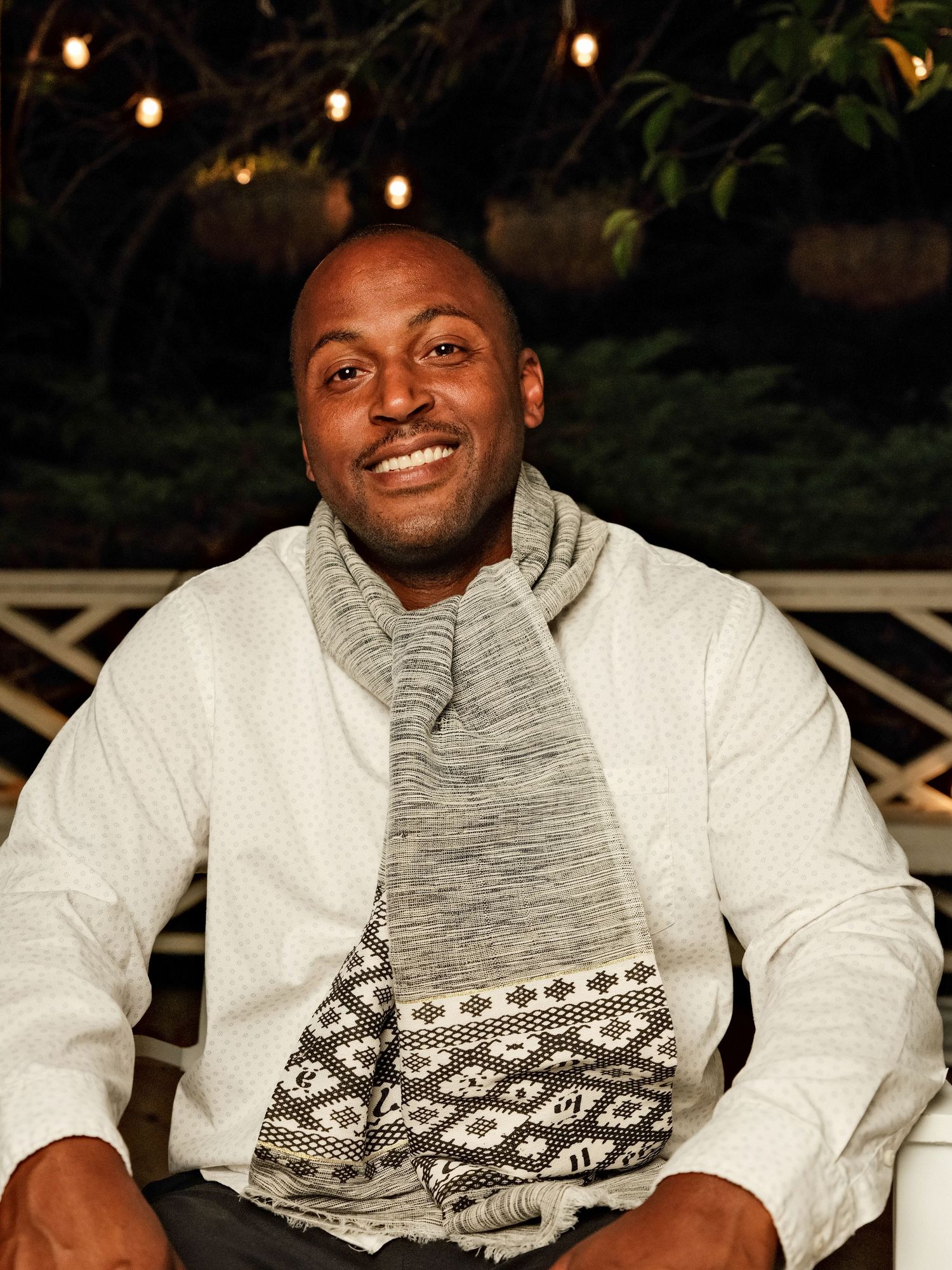Smiling man wearing a white sweater and patterned scarf seated outdoors at night with string lights behind him.