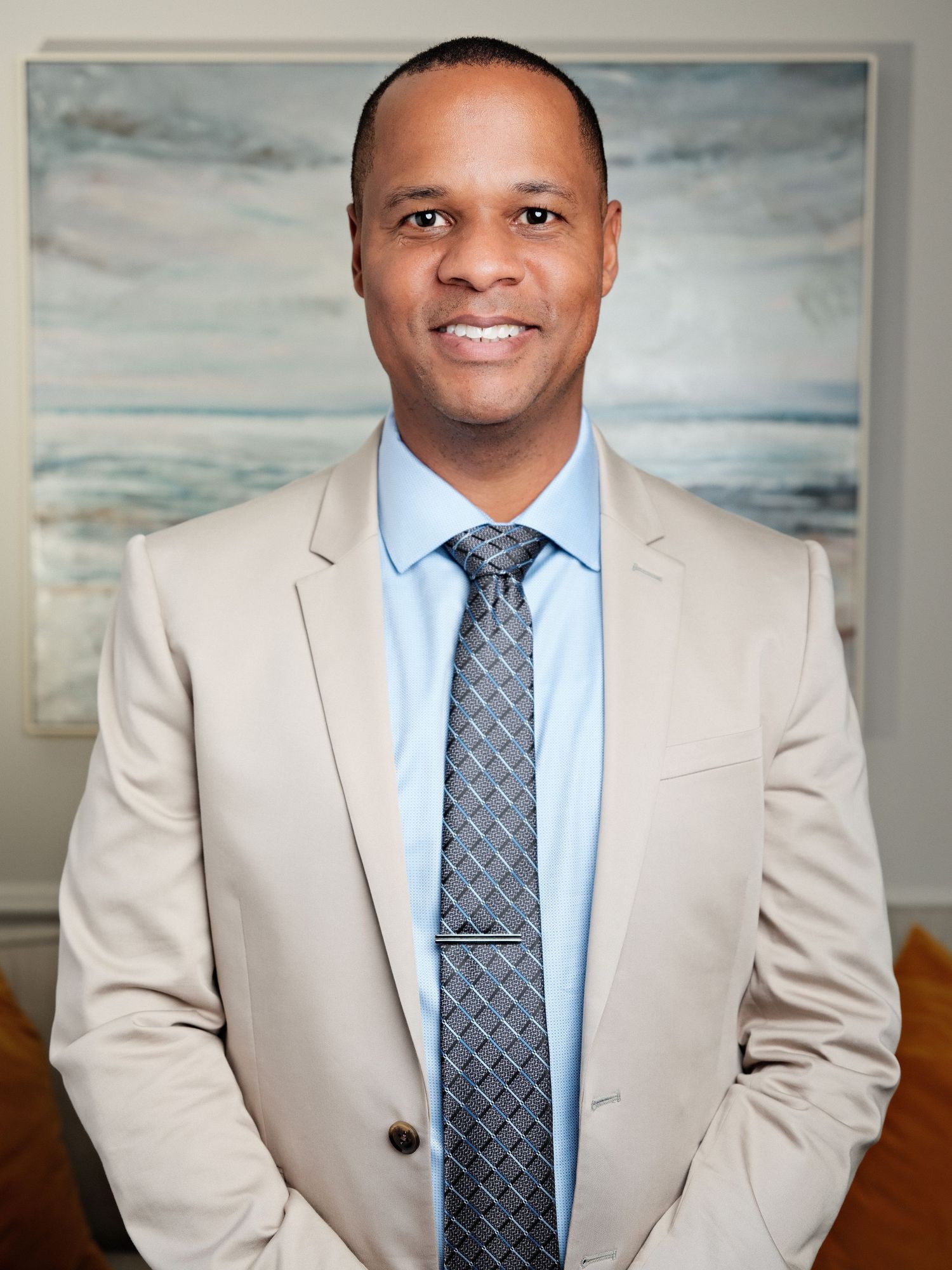 Professional man in beige suit and patterned tie smiling confidently in front of coastal artwork.