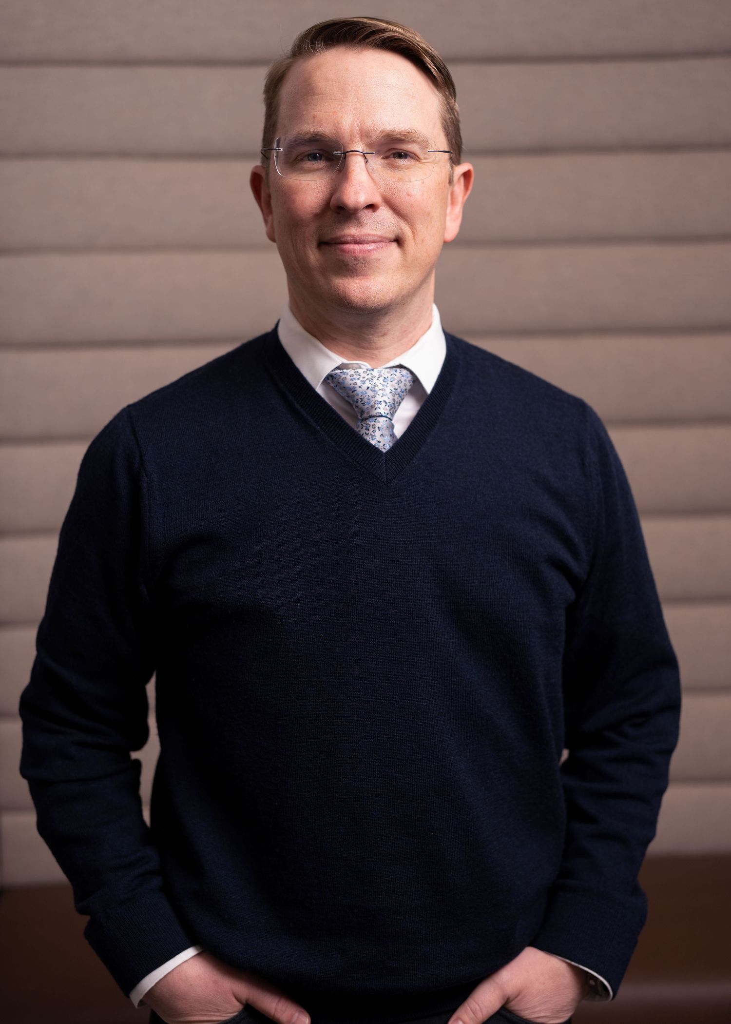 Professional man in navy sweater and tie smiling confidently in a modern office setting.