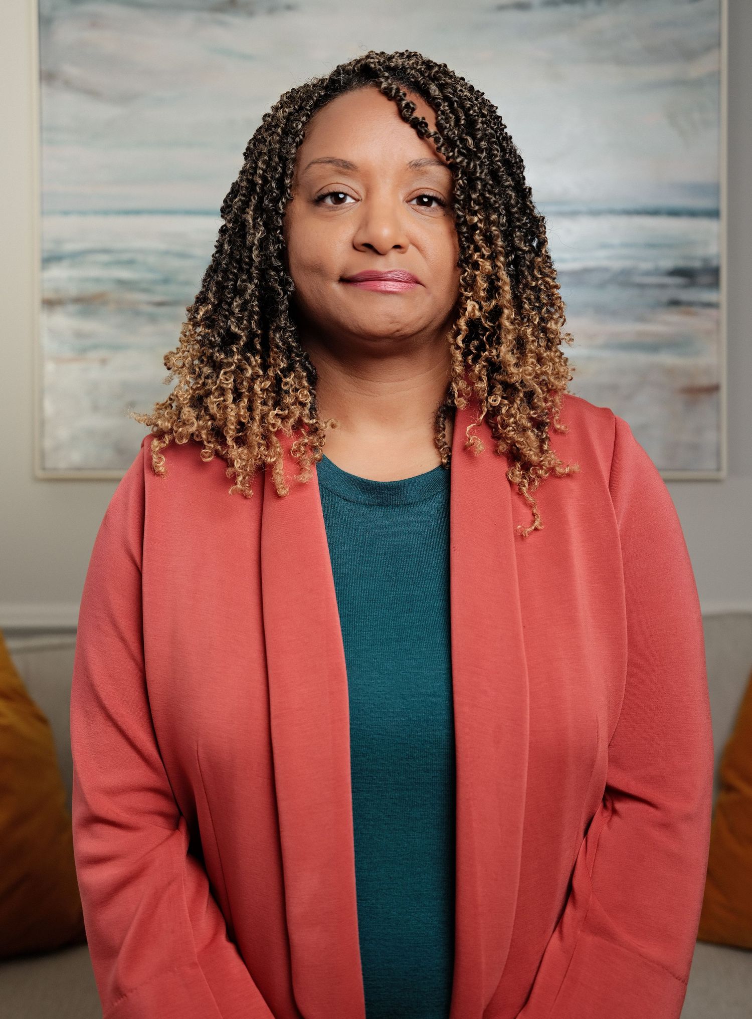 Professional woman with curly highlighted hair wearing a coral blazer over teal top, posed in a modern office setting.