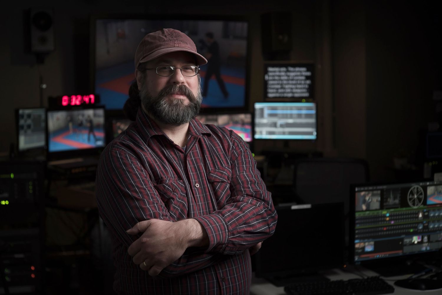 Bearded man with glasses and crossed arms stands in a dark video editing suite with multiple monitors.