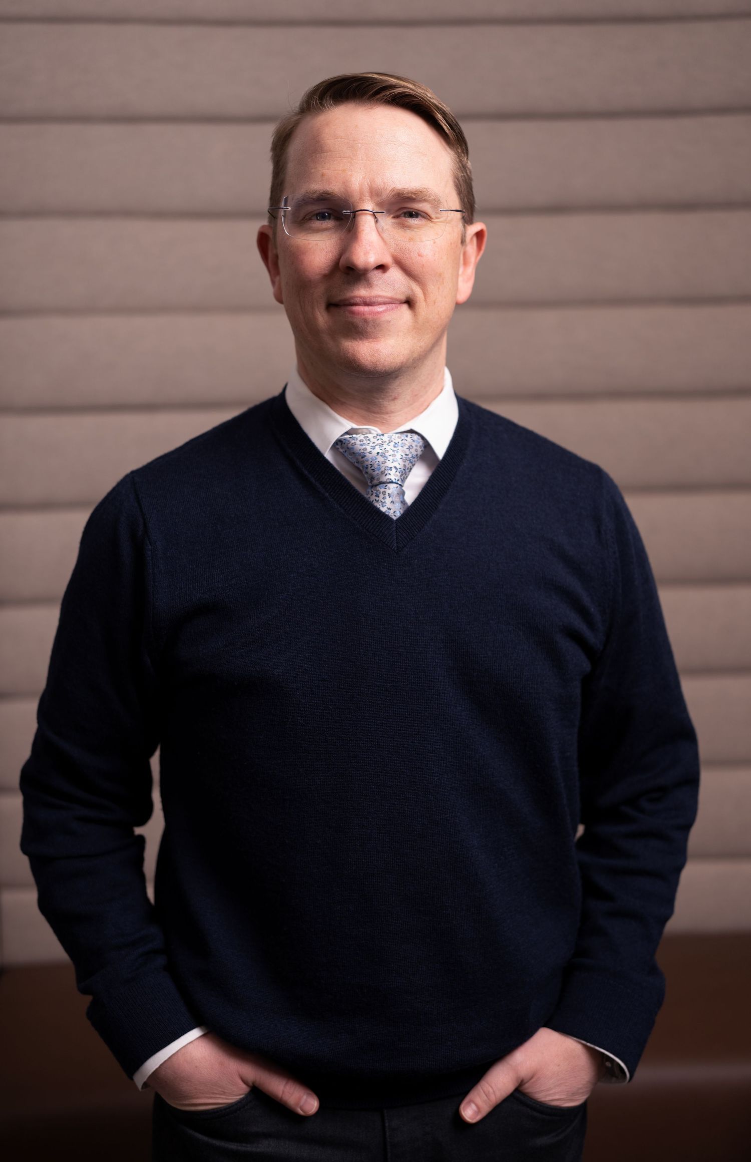 Professional man in navy sweater and tie smiling confidently in a modern office setting.