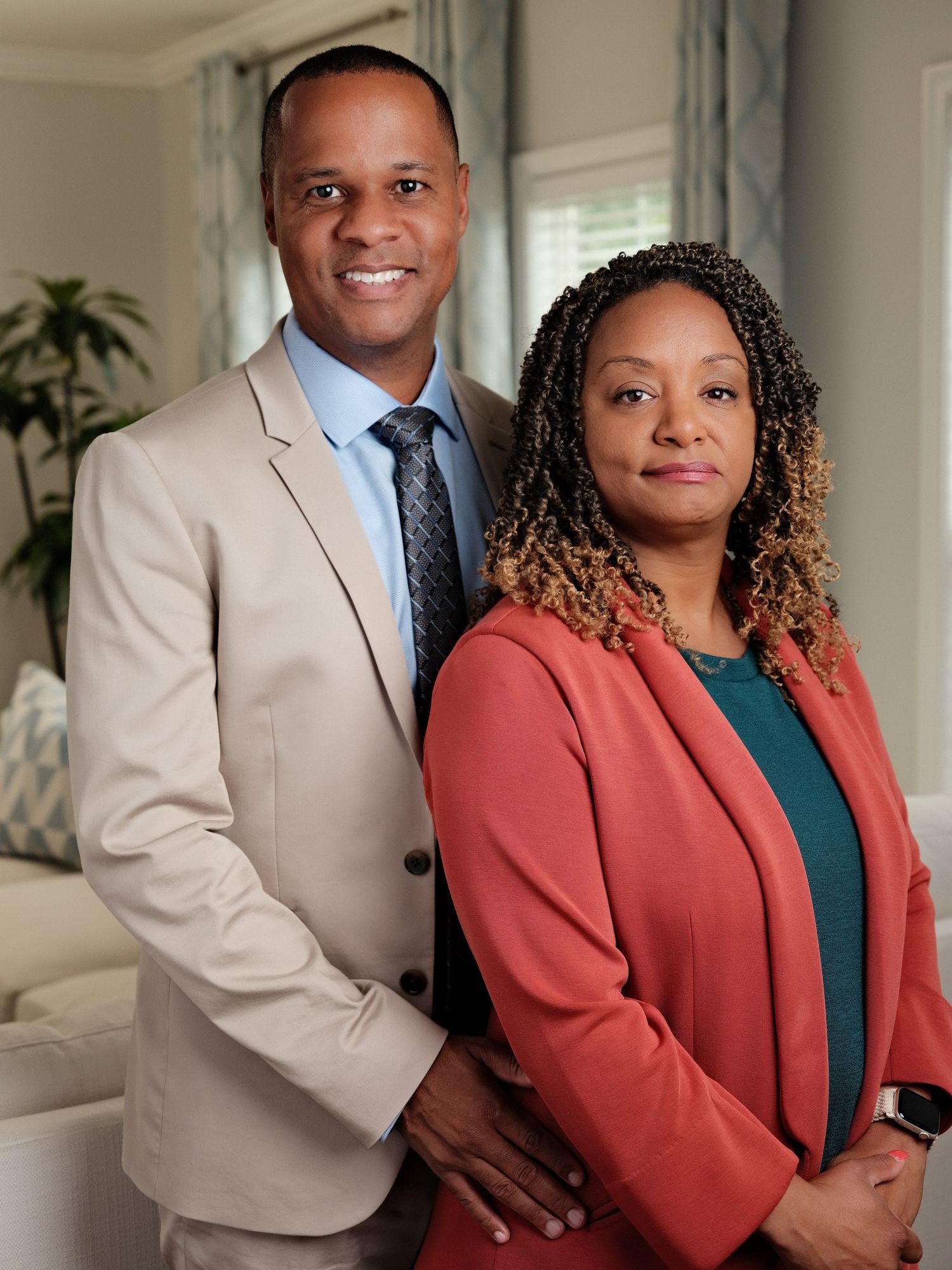 Professional couple posing indoors, man in beige suit and woman in coral blazer, smiling confidently.