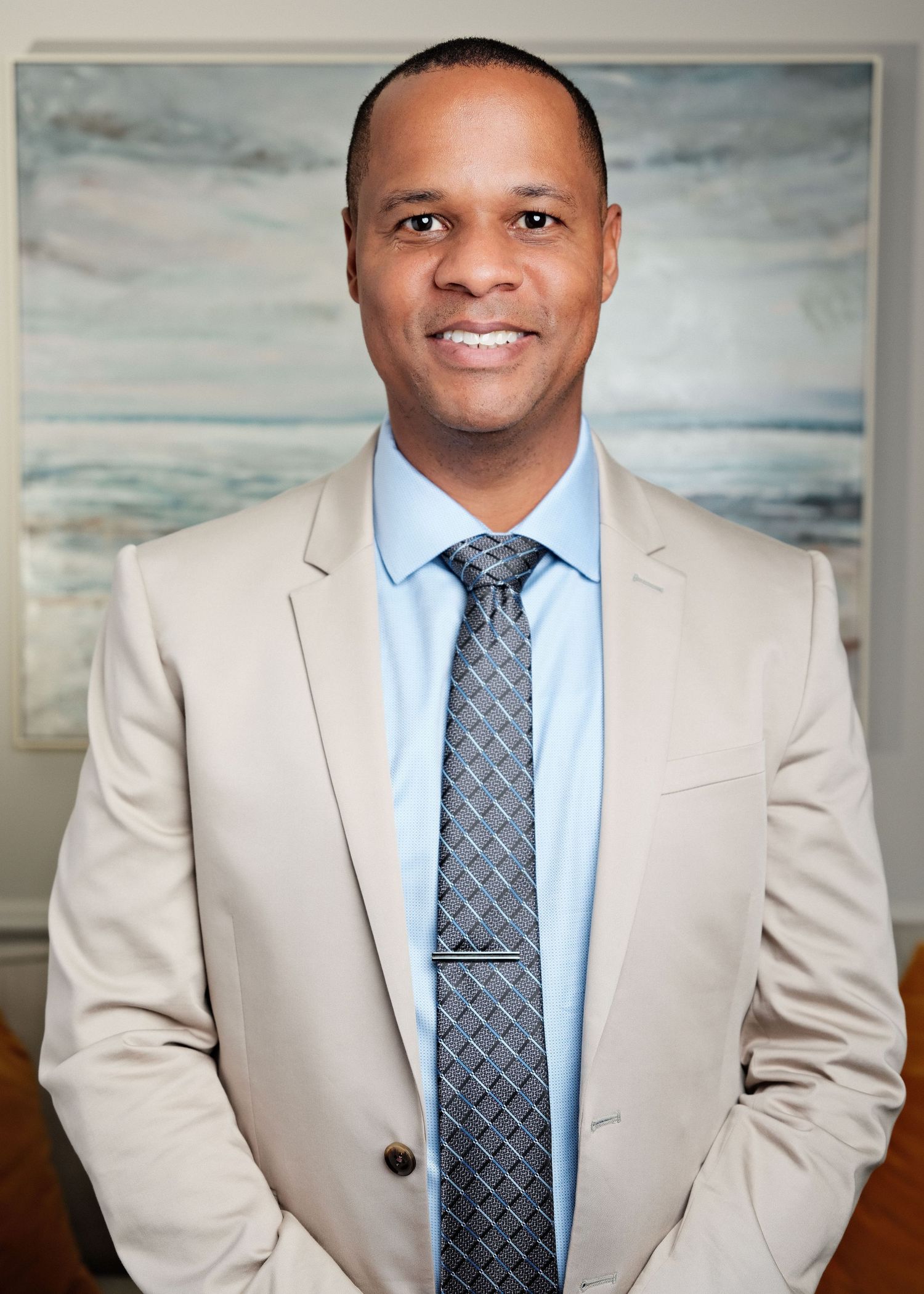 Professional man in beige suit and gray tie smiling confidently in a modern office setting.