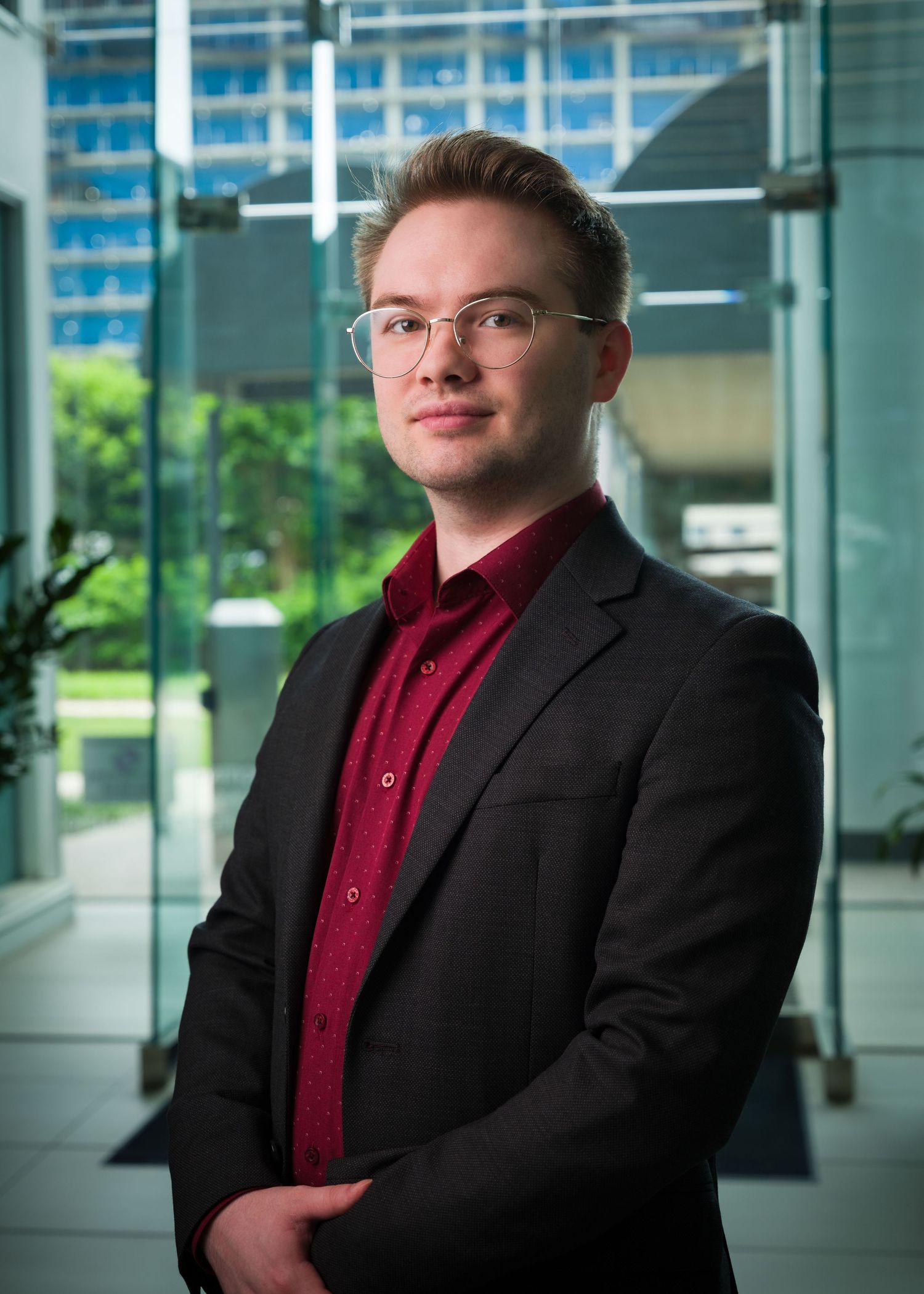 Young professional man in glasses and red shirt with black blazer posing in modern glass office building.