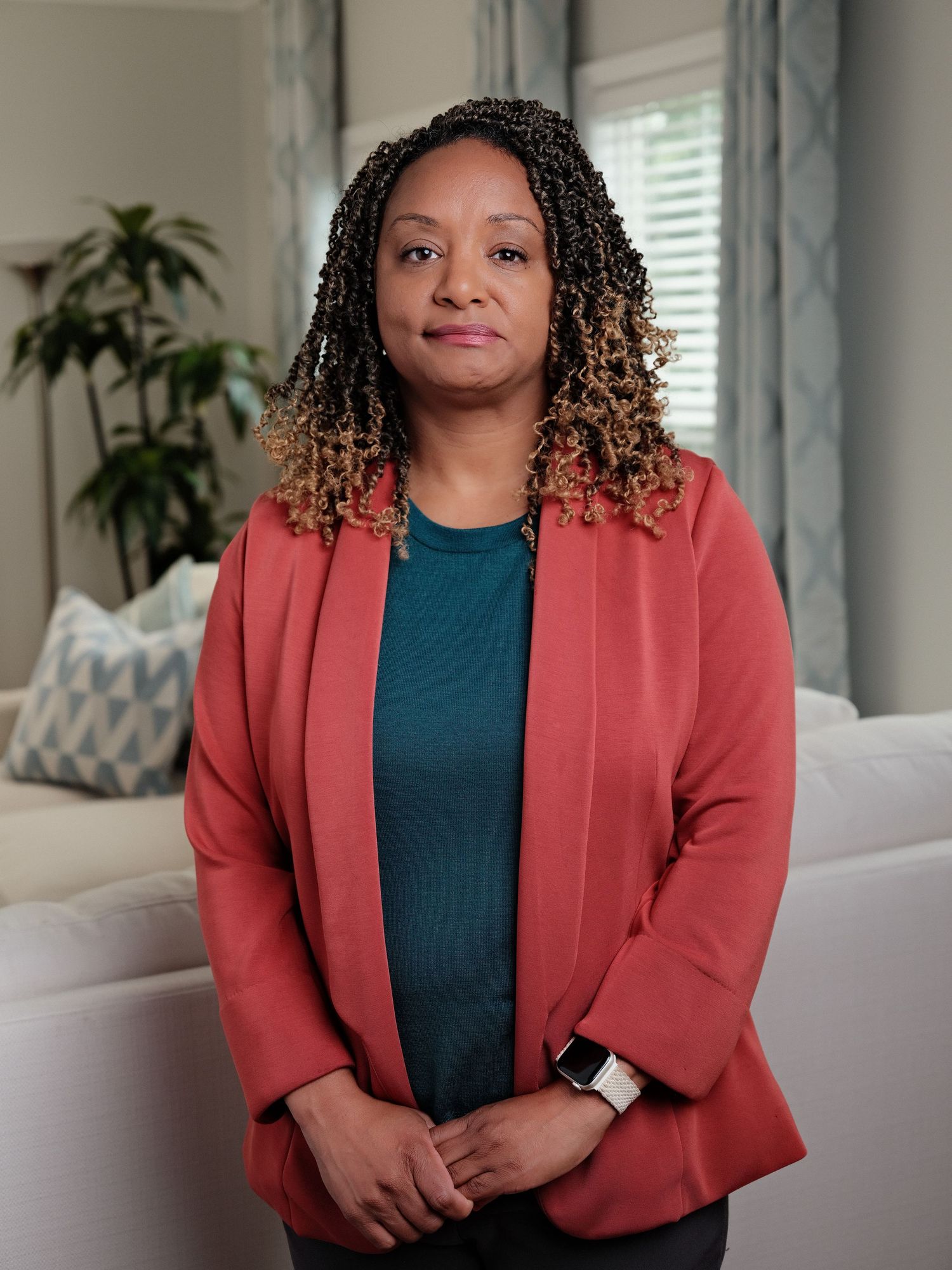 A professional woman in a coral blazer and teal top stands confidently in a modern living room setting.