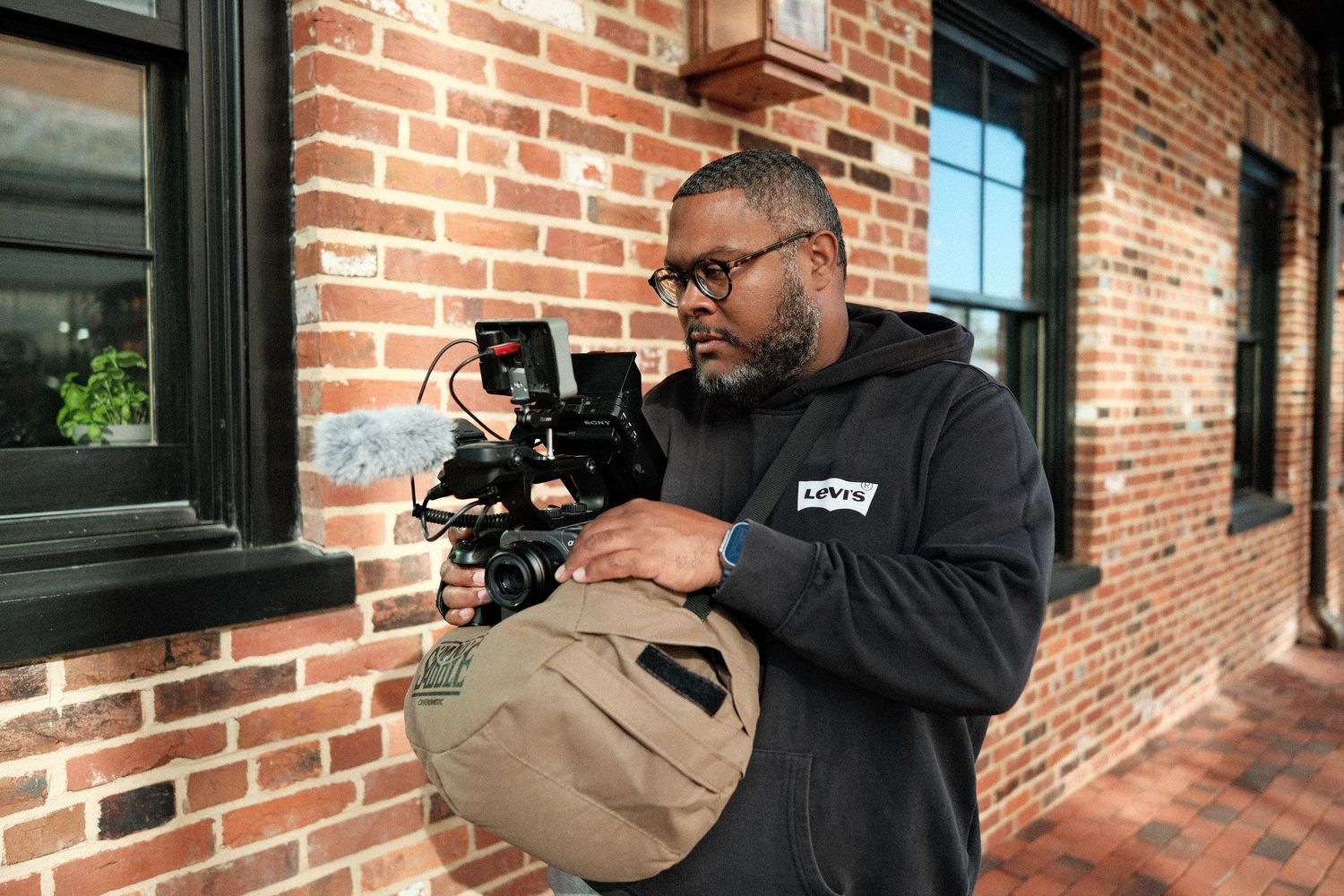 A filmmaker holds a professional cinema camera outside a brick building, wearing glasses and a dark hoodie.