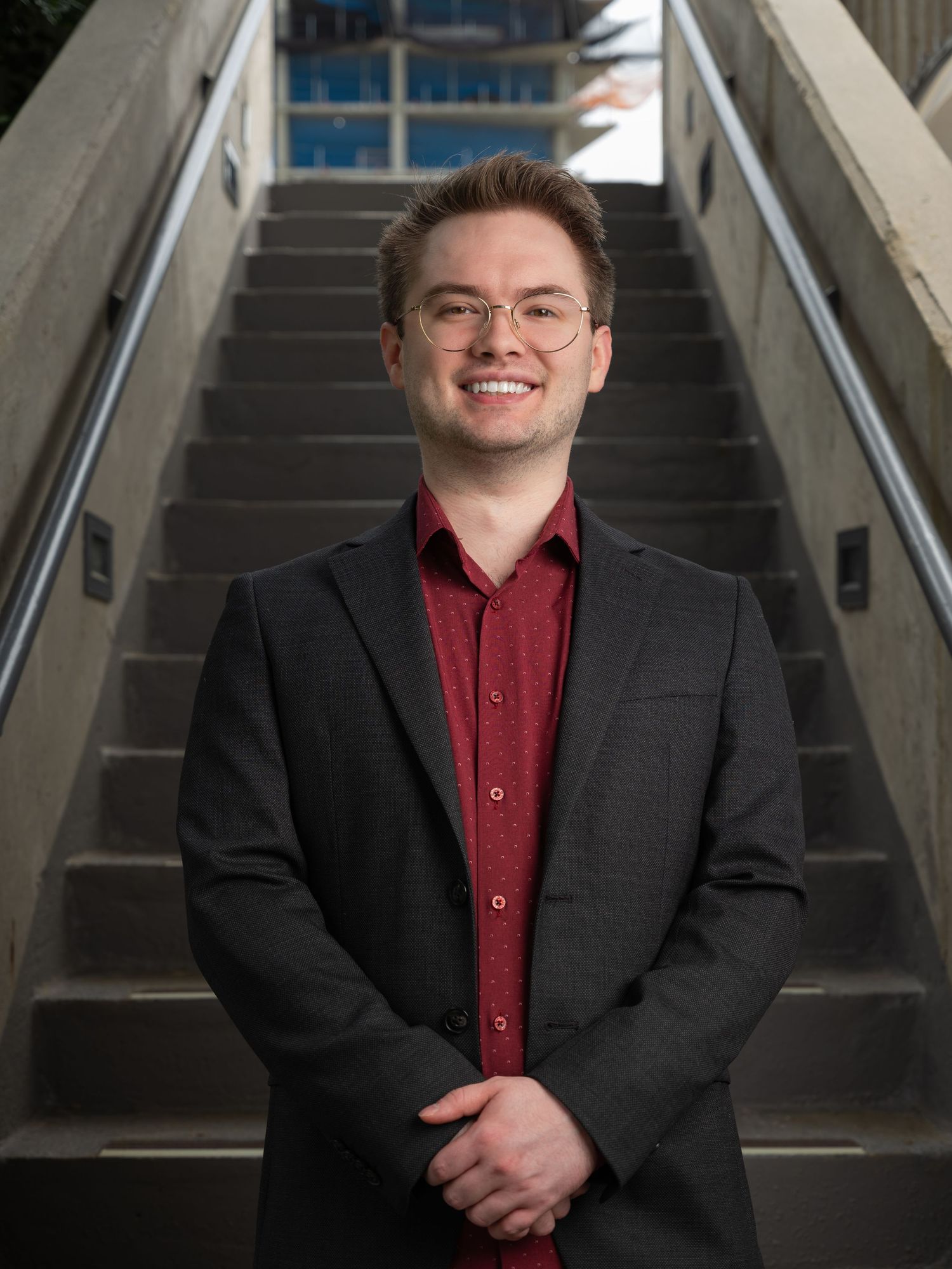Young man in glasses and red shirt with black blazer smiling on concrete staircase.
