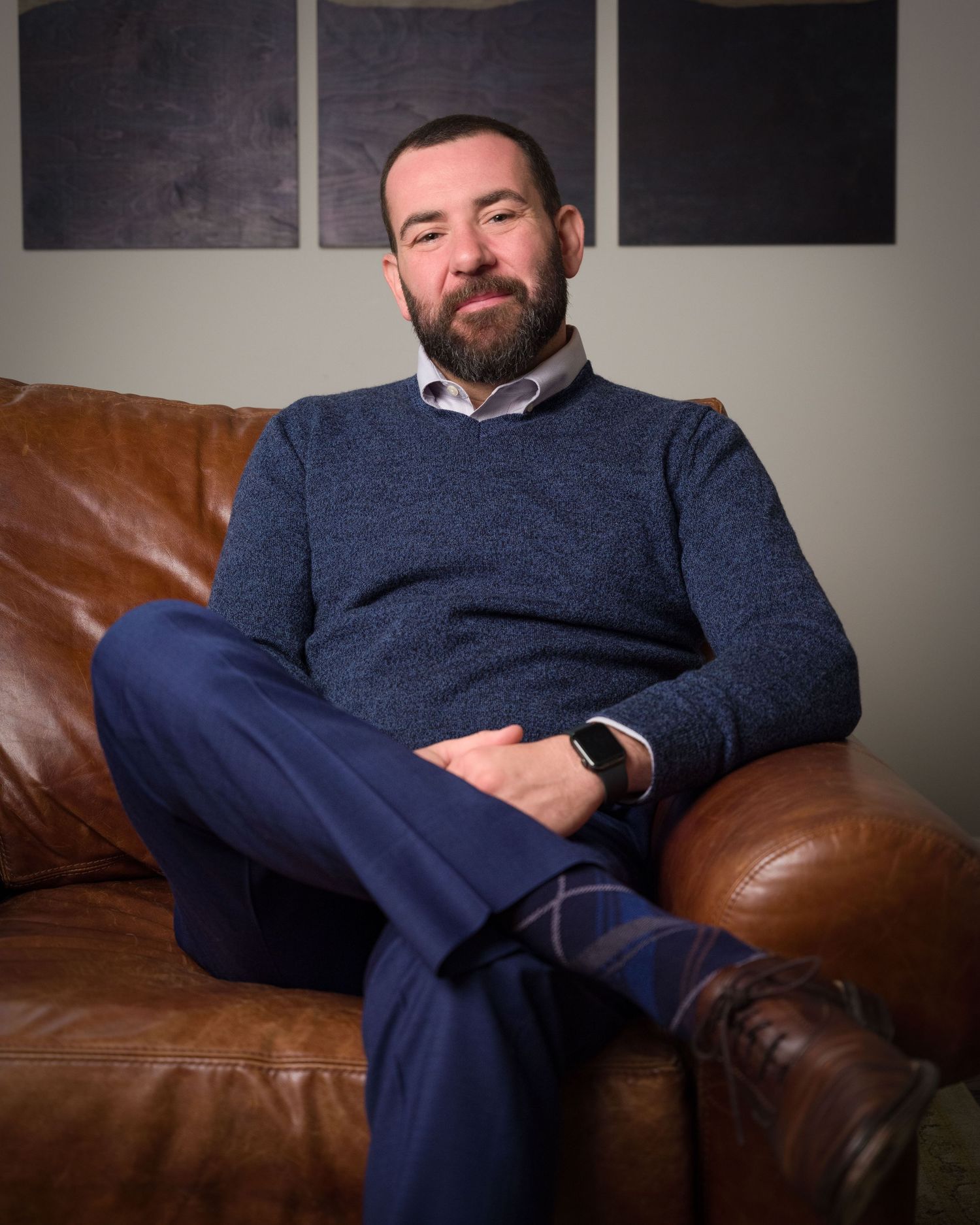 Bearded man in blue sweater and trousers sitting relaxed on brown leather sofa in modern interior.