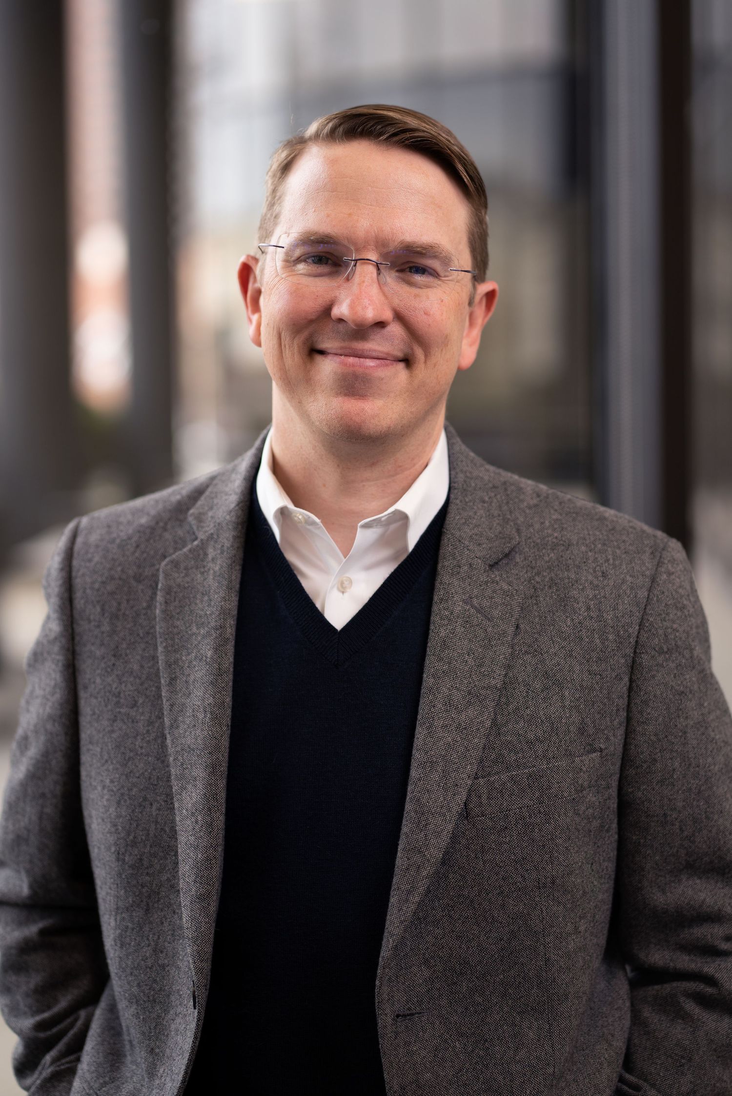 Professional man in gray blazer and glasses smiling outdoors in urban setting.