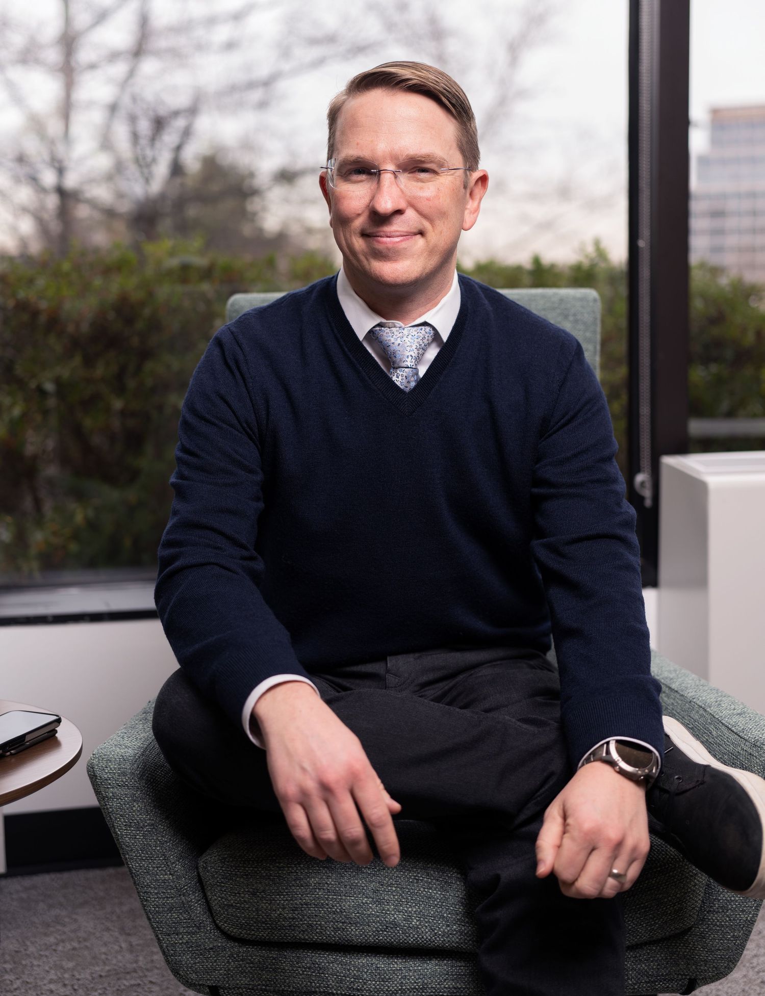 Professional man in navy sweater sitting cross-legged in modern chair, smiling confidently in bright office setting.