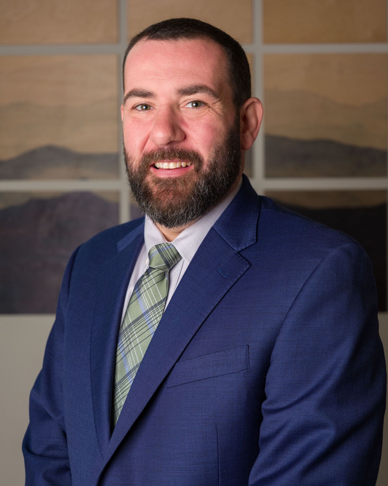 Professional headshot of a smiling man with a beard wearing a navy blue suit and green plaid tie.