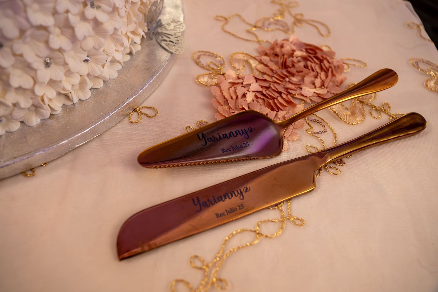 Personalized gold wedding cake server and knife set engraved with names, displayed beside a white floral wedding cake. Sweet 16/Quinceañera