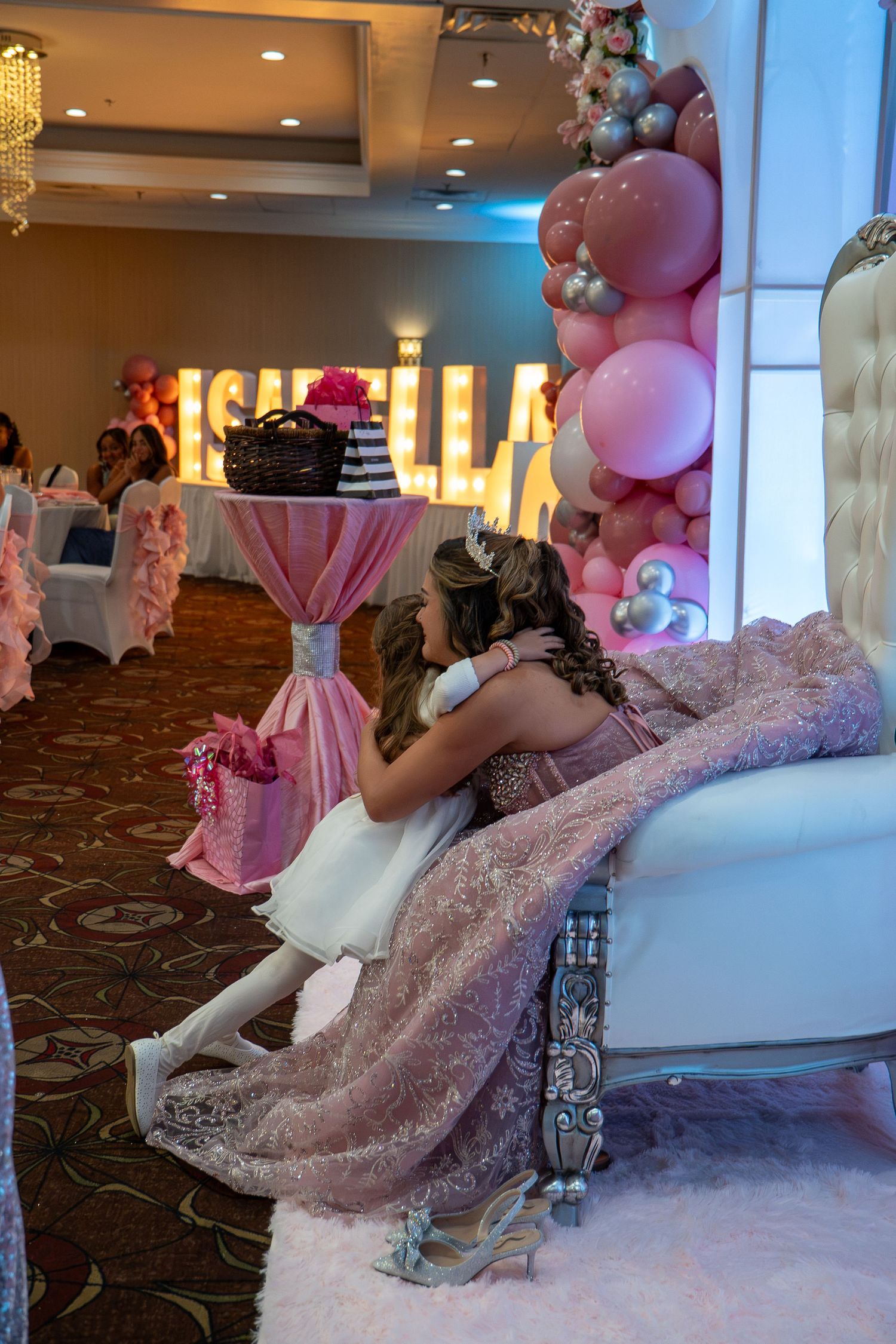 Two girls in pink dresses embrace on a white throne chair at Isabella's quinceañera party with balloon decorations. Sweet 16!