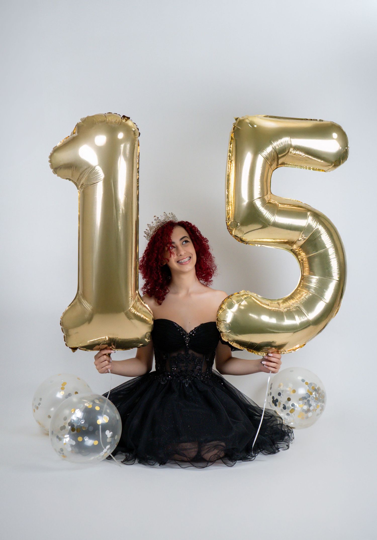 Young woman in black dress celebrating 15th birthday with gold number balloons and confetti balloons.