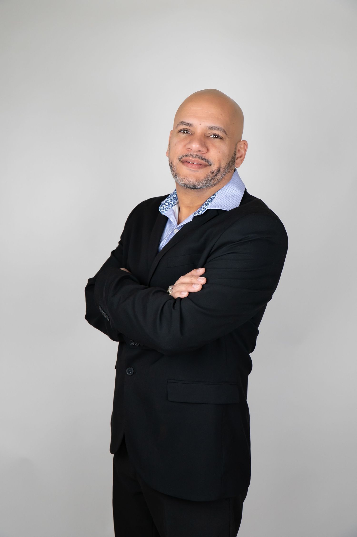 Confident bald man in black suit with arms crossed smiling against gray background.