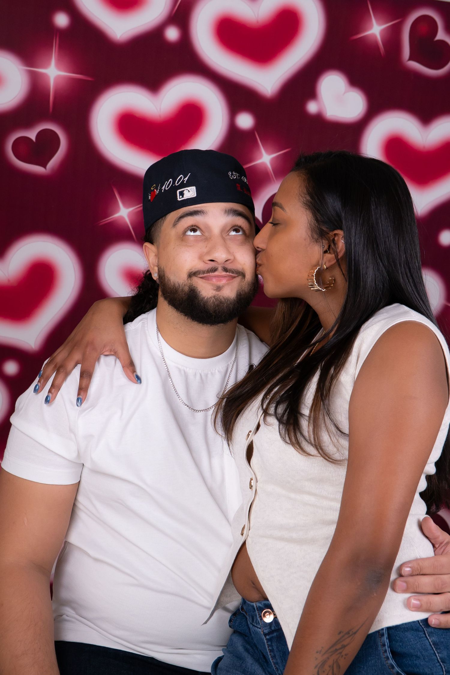 Couple posing for a Valentine's Day photo with heart backdrop, woman kissing man on cheek.