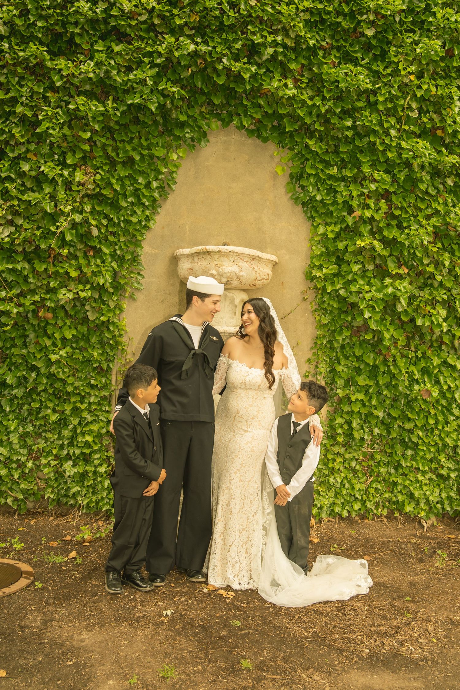 Navy sailor groom and bride in lace dress pose with two boys in black suits before ivy-covered garden wall.