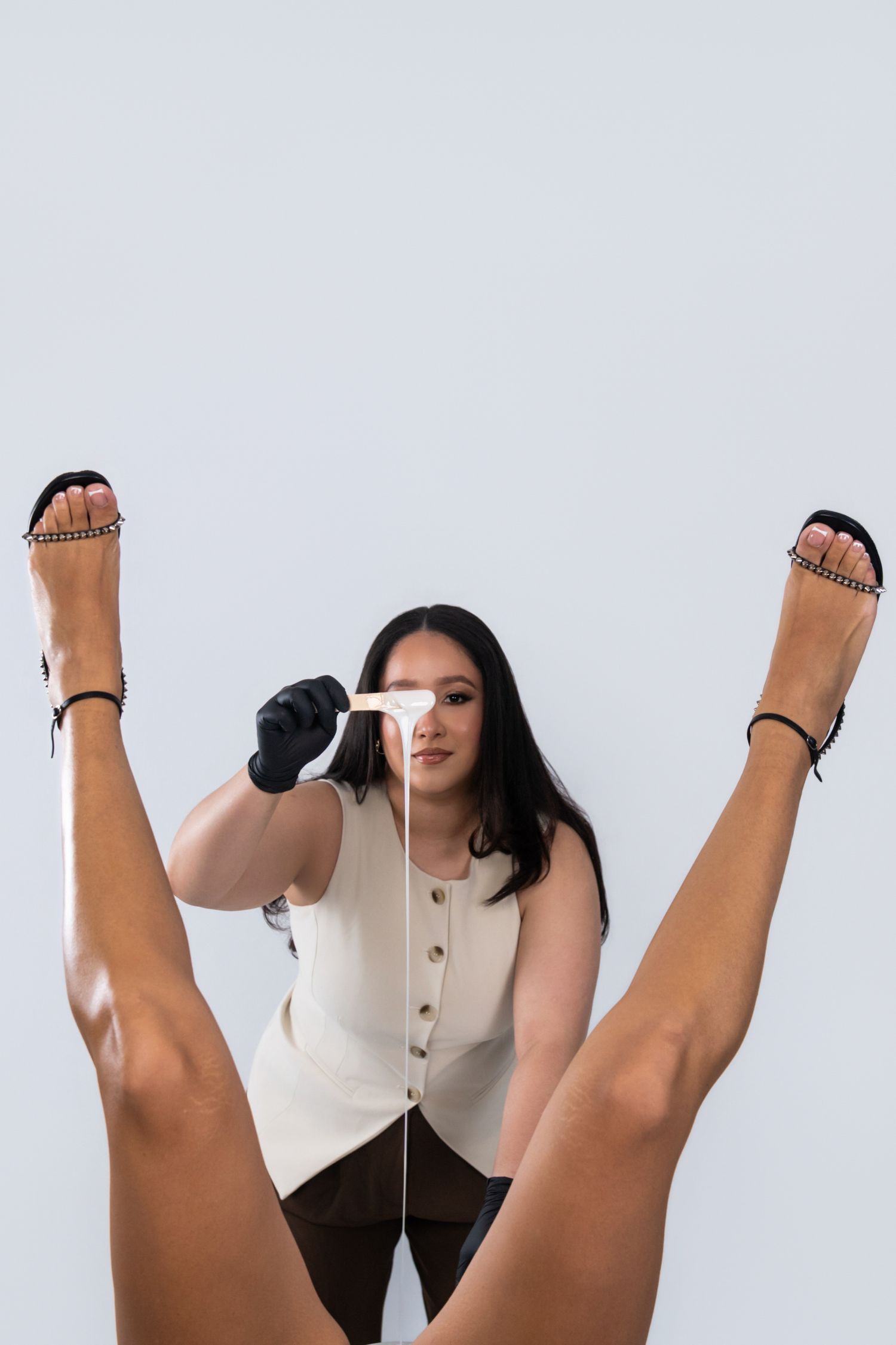 Esthetician applying wax strip for hair removal on raised legs of client wearing black heeled sandals.