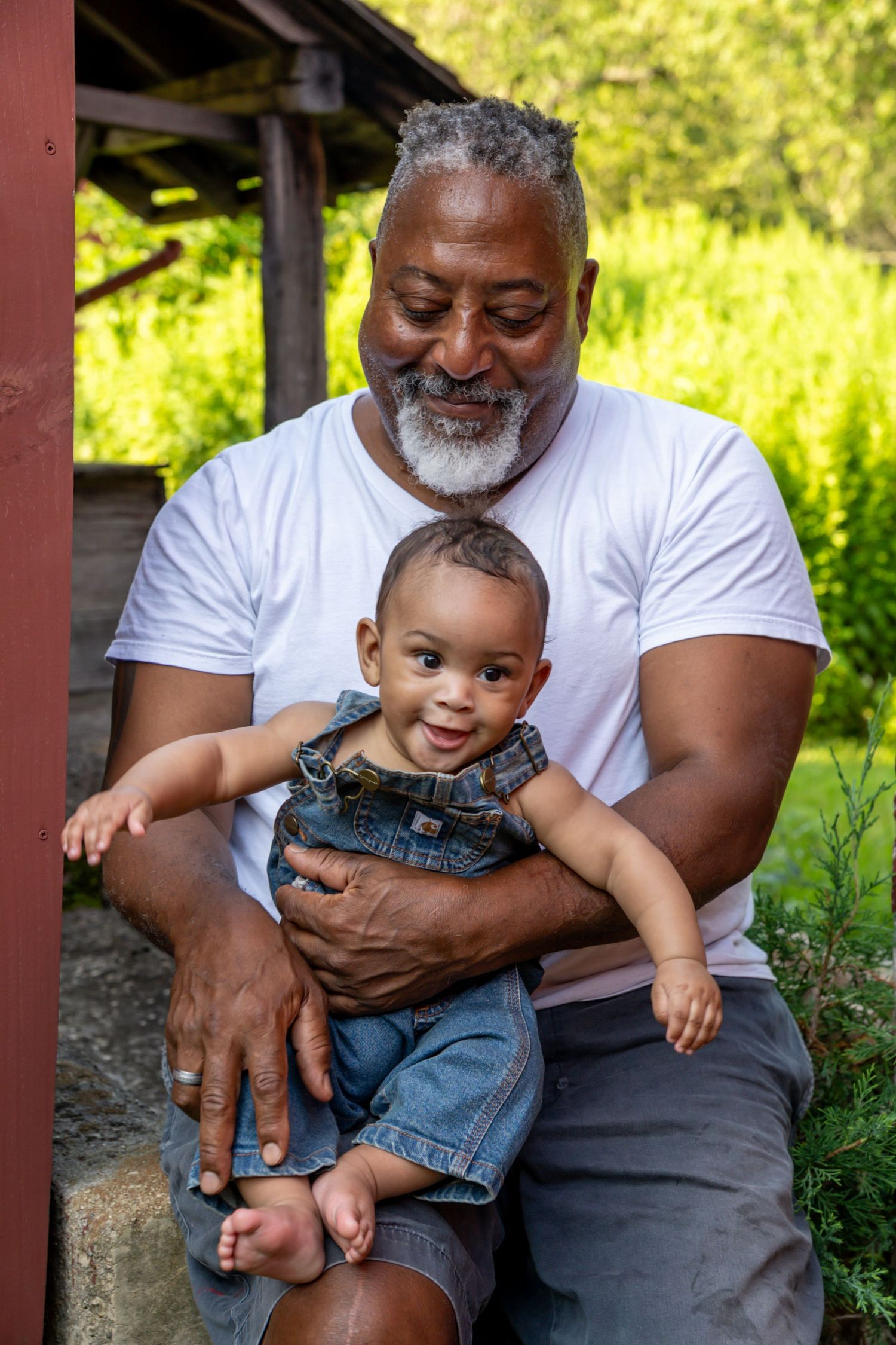A grandfather smiles warmly while holding his baby grandchild in denim overalls outdoors.