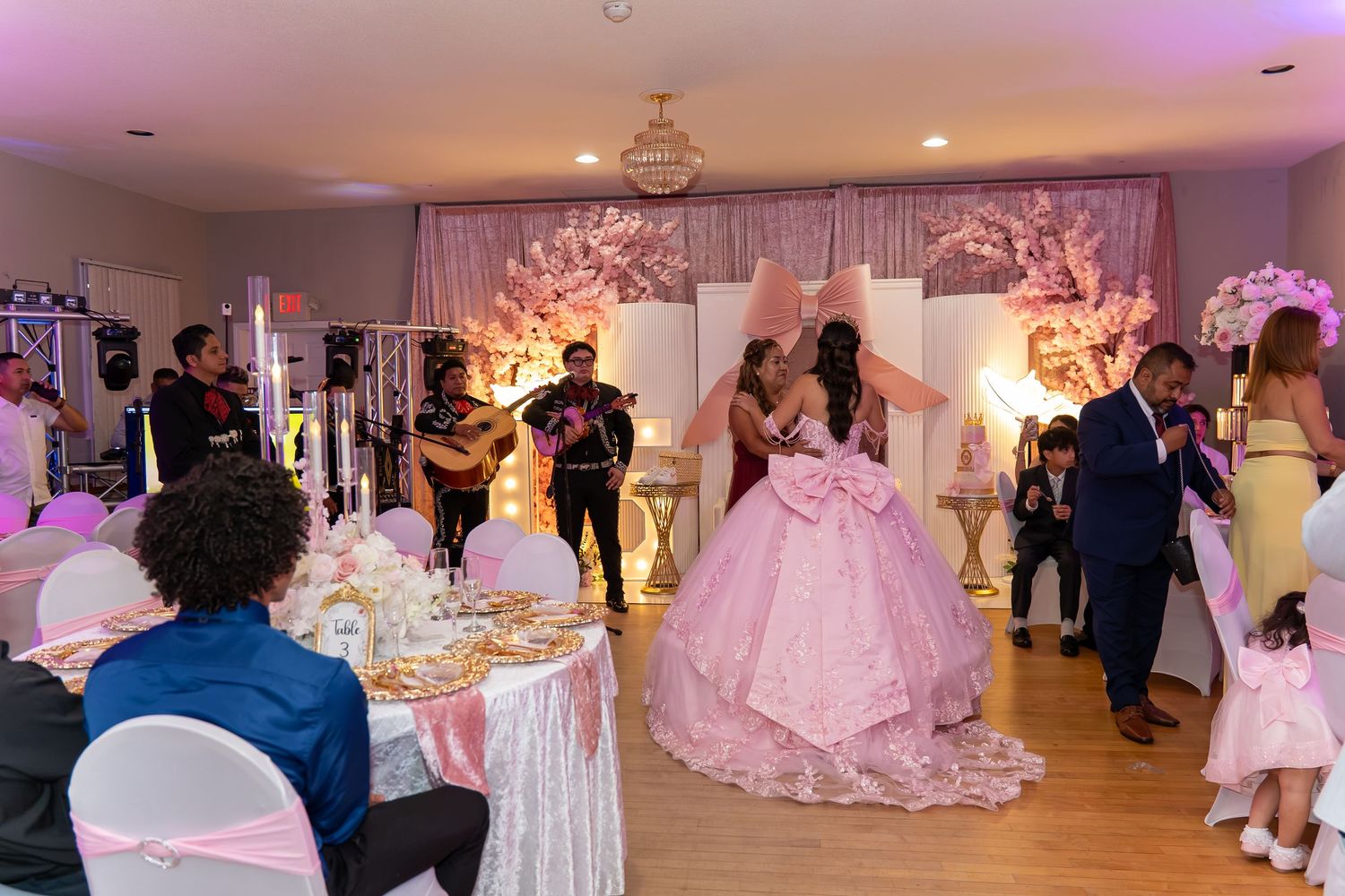 Elegant quinceañera celebration with mariachi band, pink ballgown, floral decor, and guests in a decorated banquet hall.