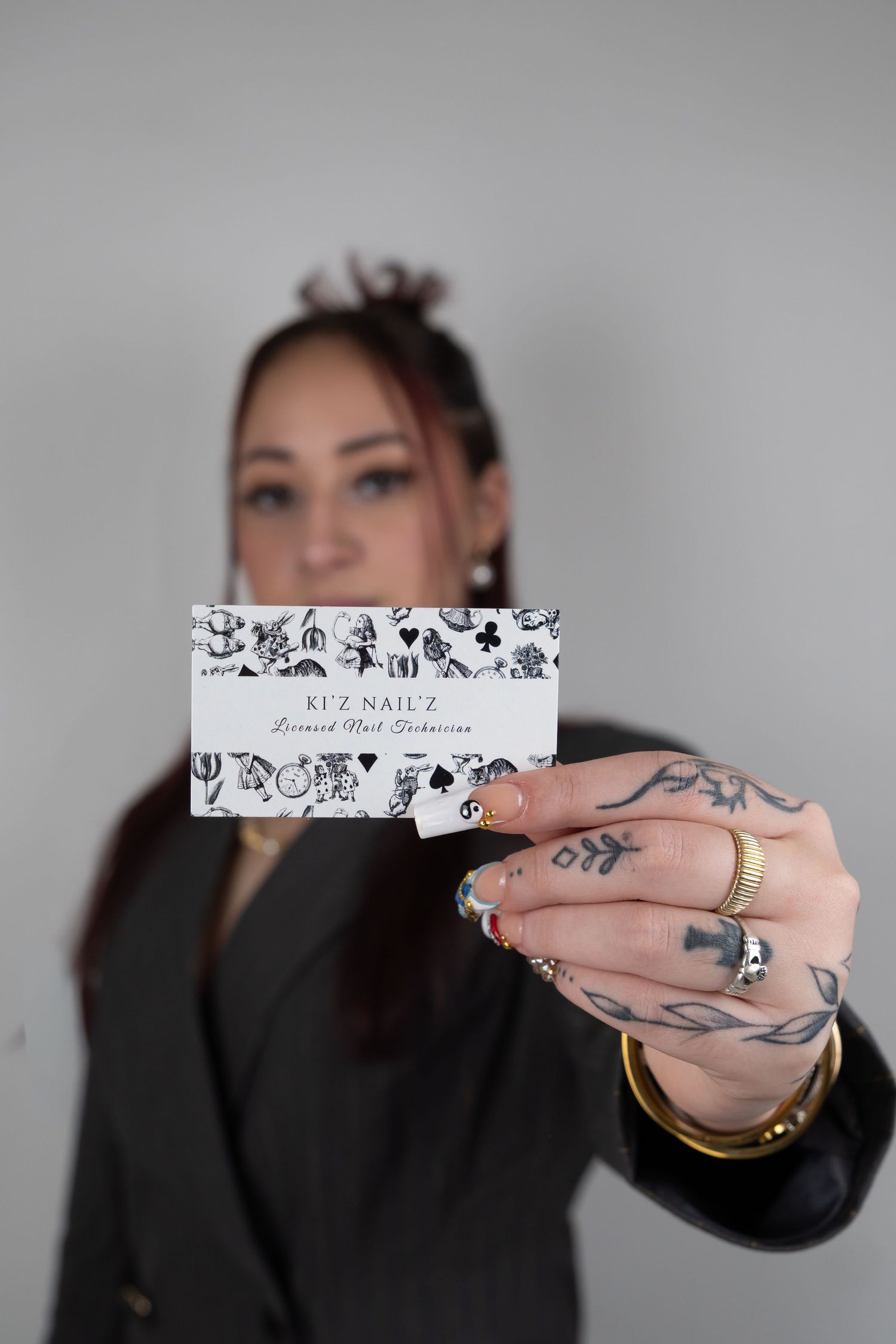 Tattooed nail technician holding Kez Nailz business card with decorated nails and gold rings.