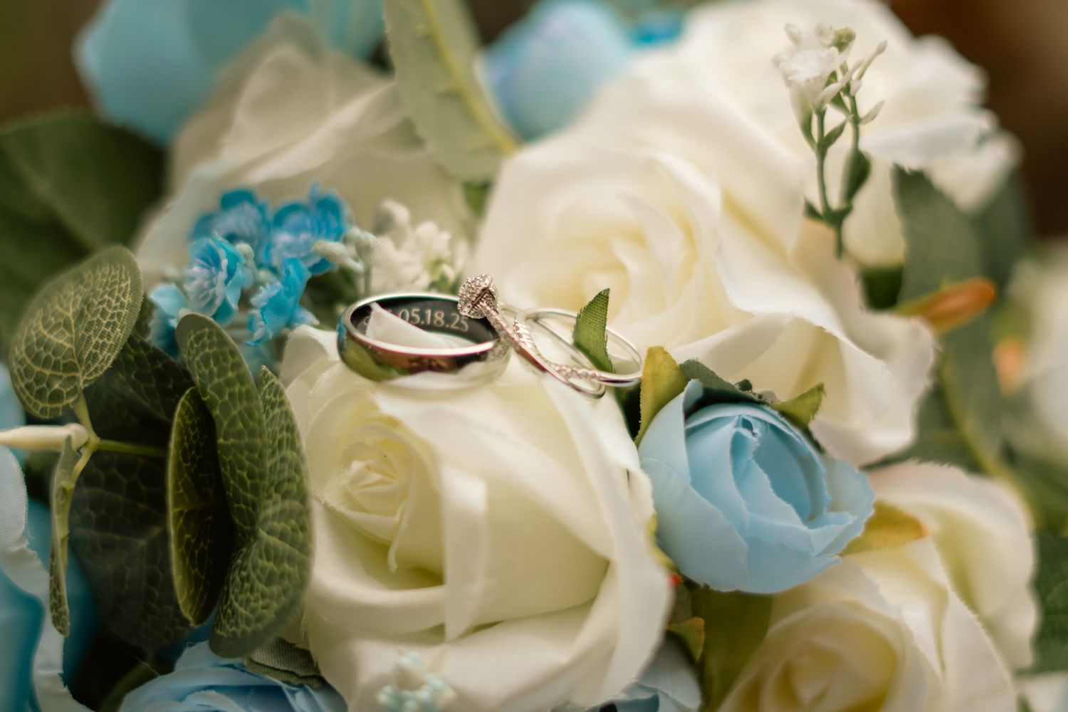Two wedding rings rest elegantly on a bouquet of white and blue roses with green foliage.