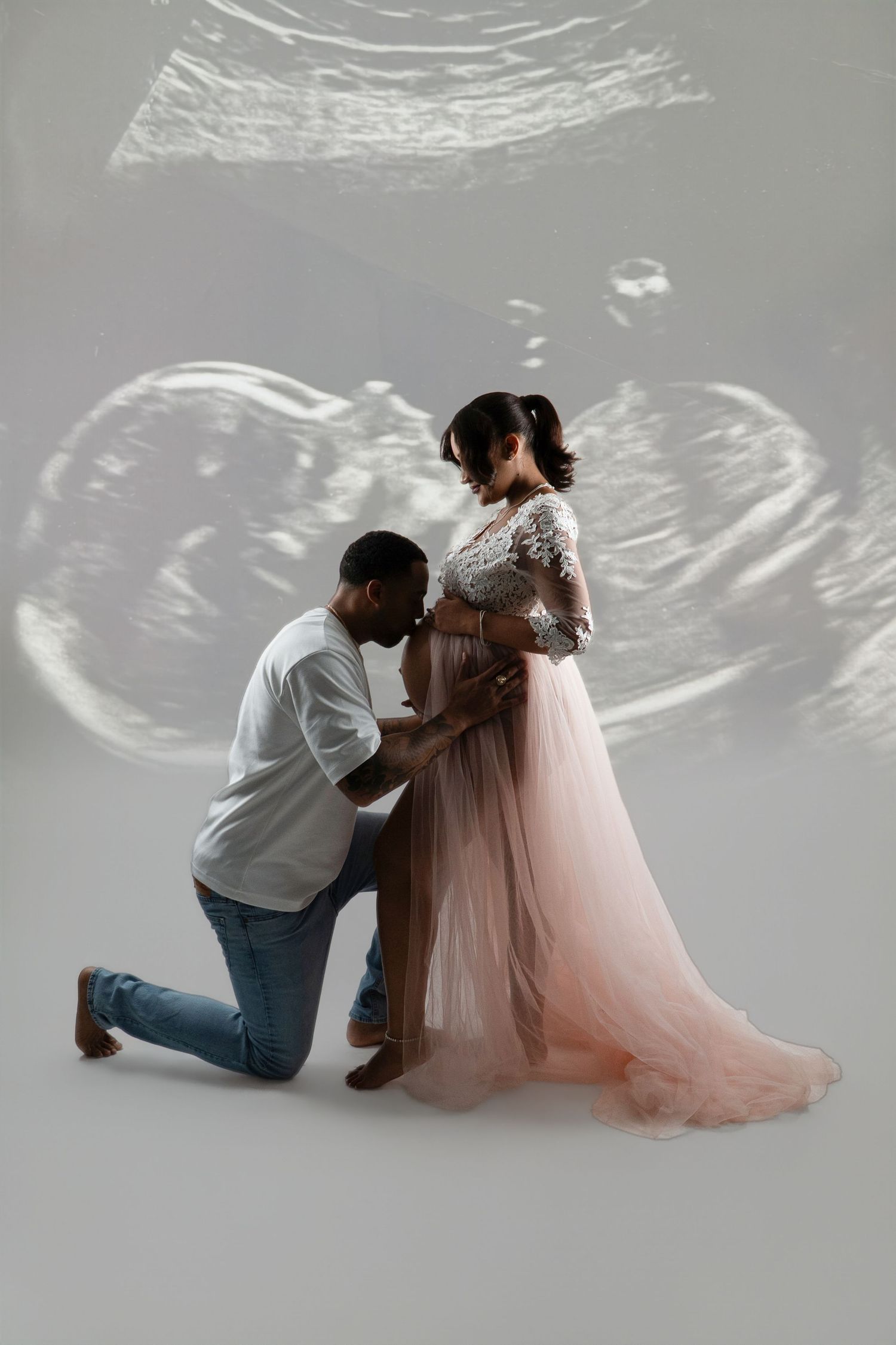 Man kneeling to kiss the baby bump of a pregnant woman in a pink tulle maternity gown against a backdrop of projected ultrasound images.