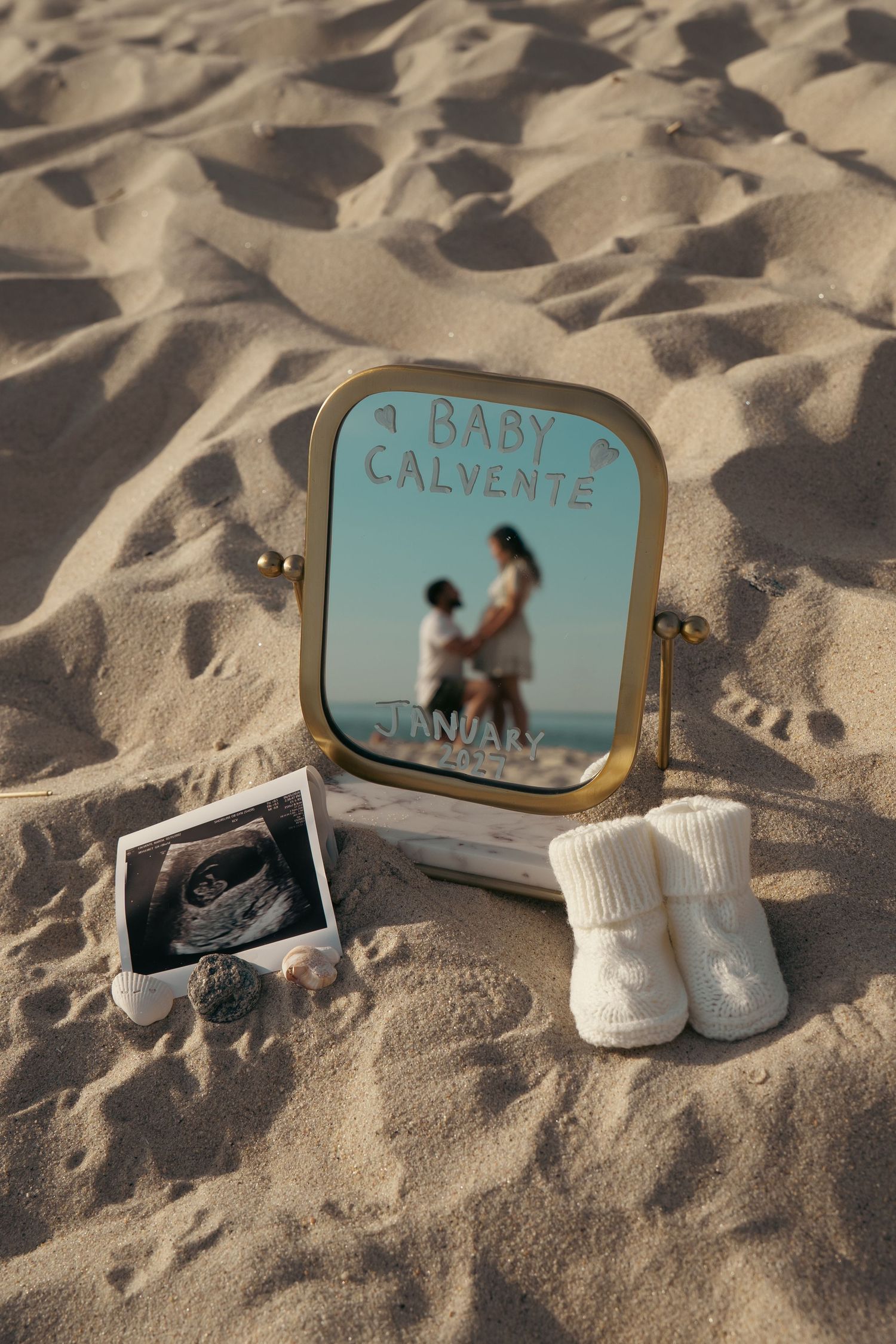 Beach pregnancy announcement with mirror, ultrasound photo, baby booties, and 'Baby Calvente January' written on sand.