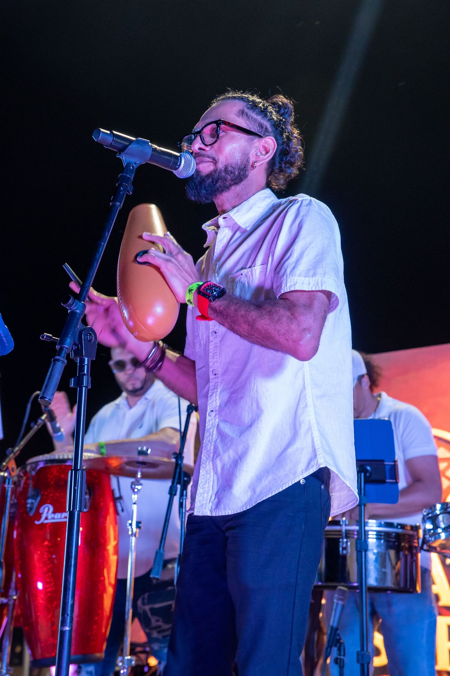Bearded musician with glasses plays a gourd instrument at a microphone stand during a live percussion band performance.