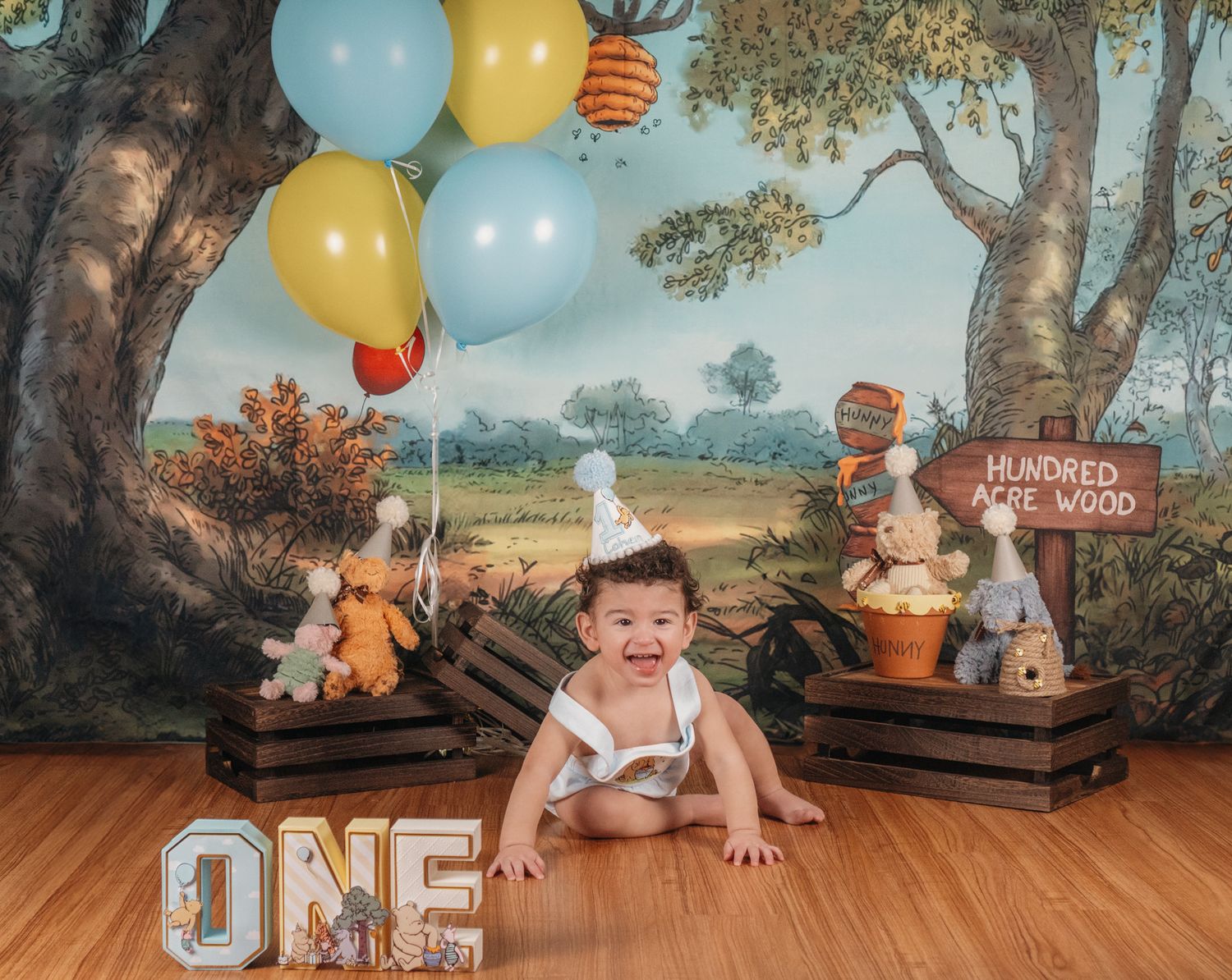 Happy baby celebrates first birthday with Winnie the Pooh themed balloons and woodland backdrop.