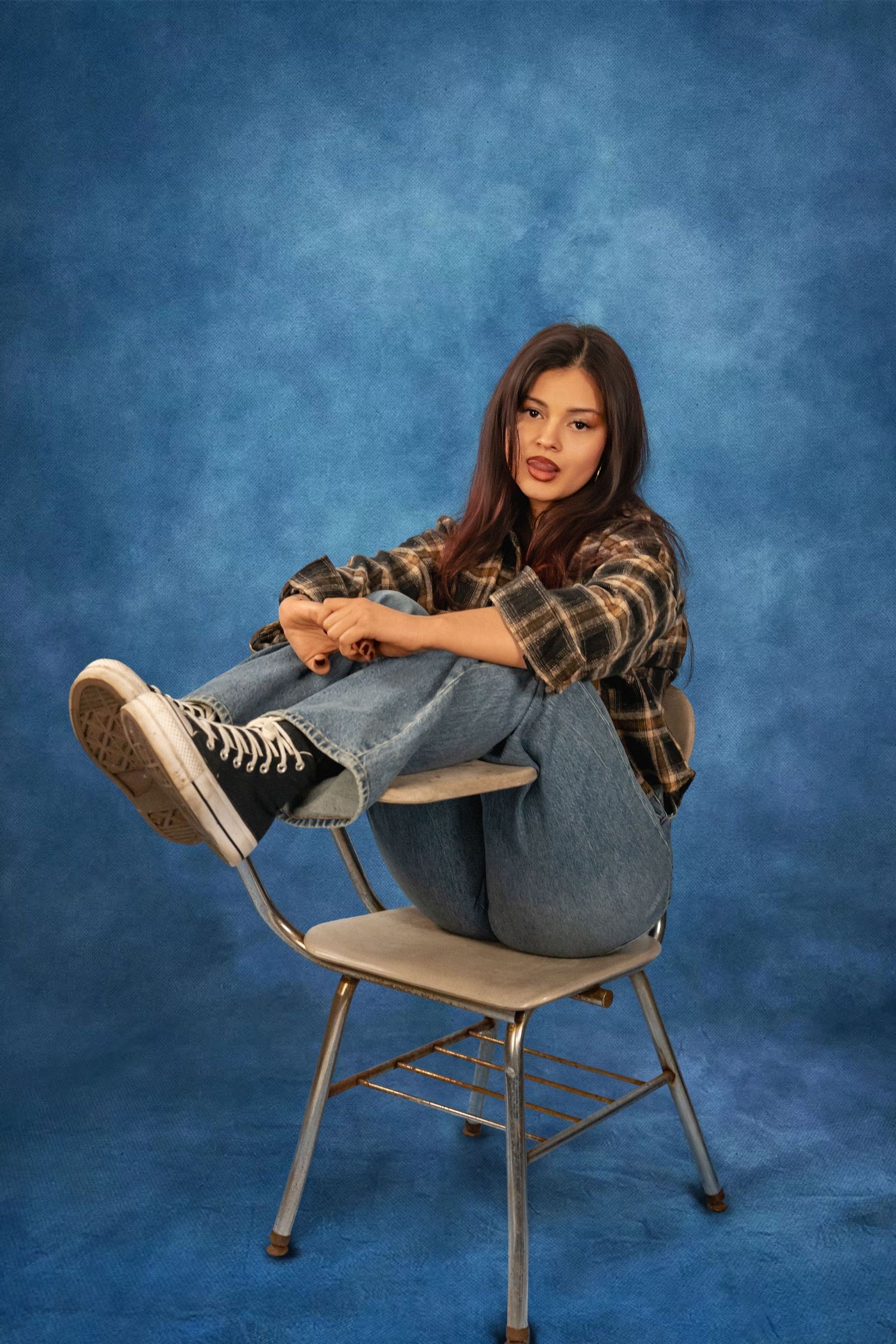 Young woman with long dark hair in a plaid flannel shirt and jeans sits in a school desk chair, feet up, against a blue studio backdrop.