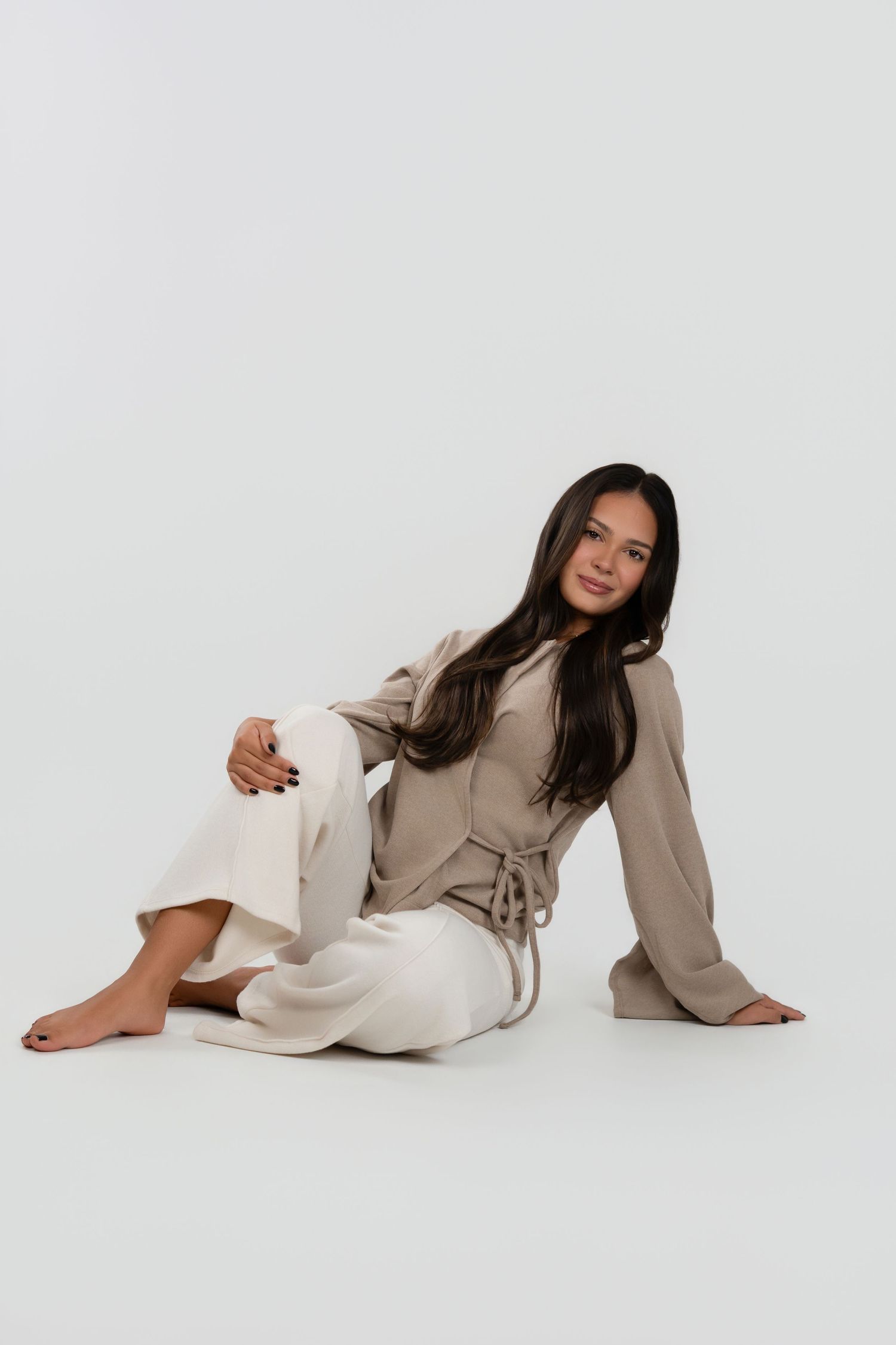 Woman with long dark hair sitting barefoot on a white studio floor, wearing a beige wrap-style top and cream wide-leg pants.