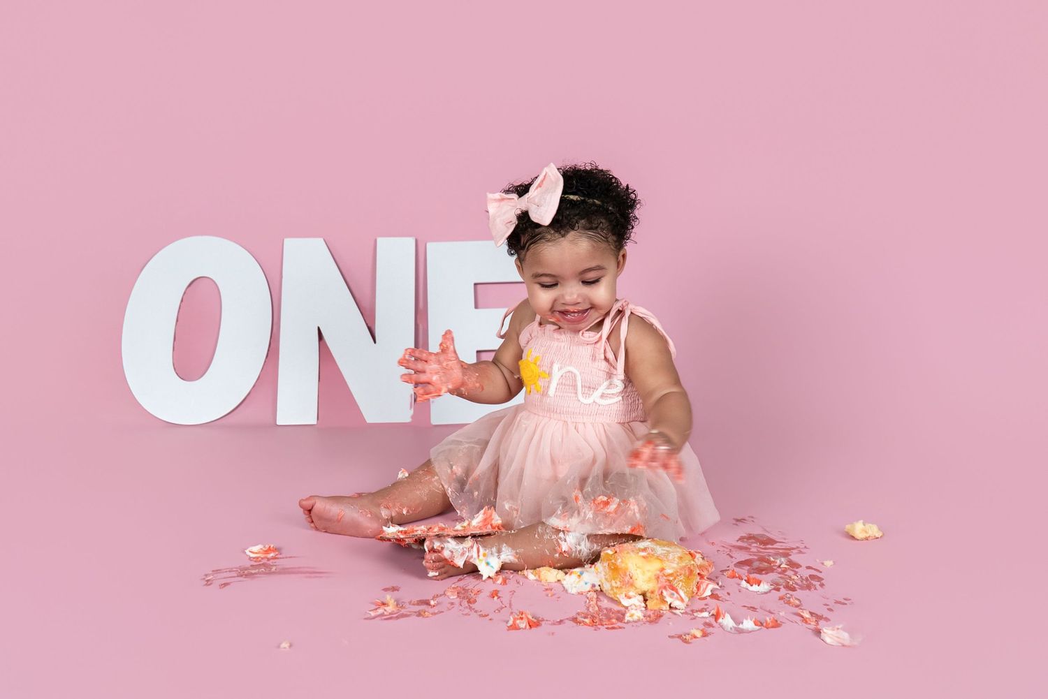 Baby girl in a pink tutu dress and bow sits smiling beside a large ONE sign during a first birthday cake smash photoshoot.