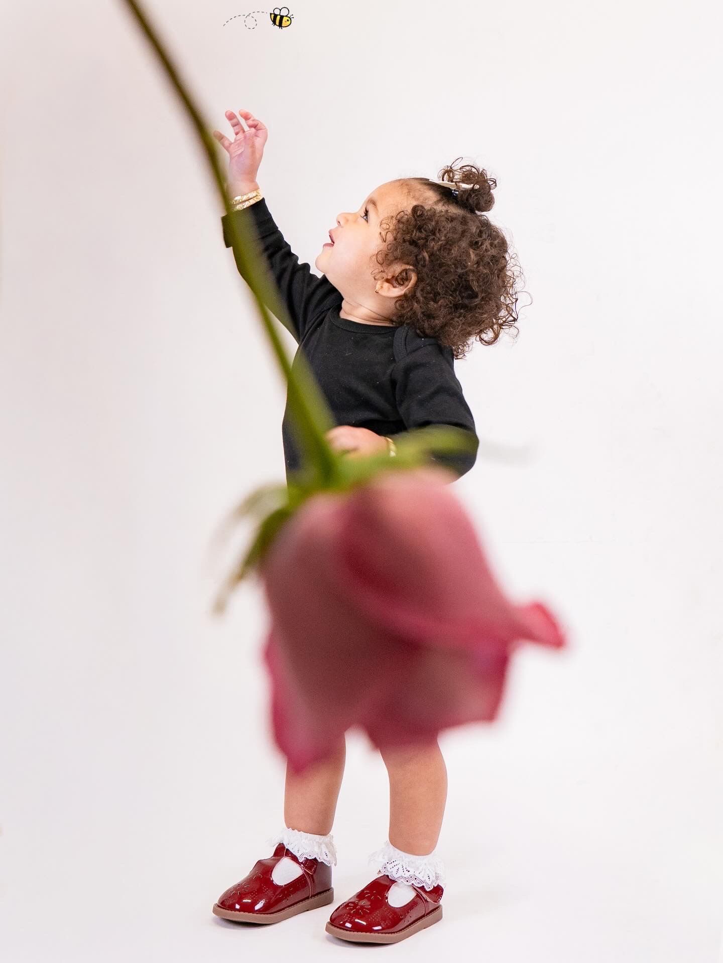 A curly-haired toddler in a black outfit and red shoes reaches up toward a bee, with a blurred pink rose in the foreground.