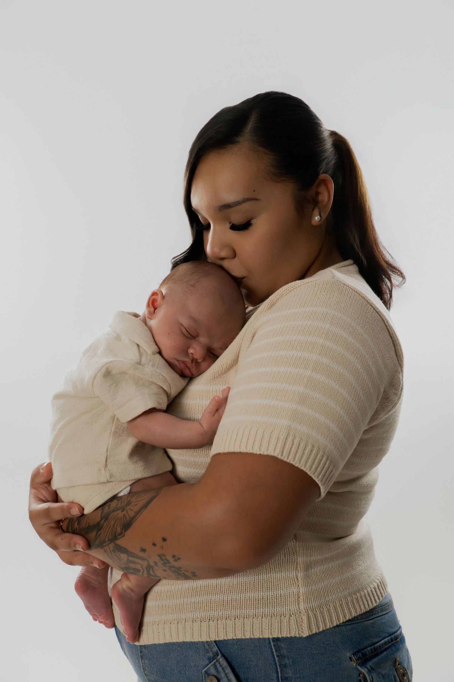 A mother tenderly kisses her sleeping newborn baby while holding them close against a white background. Newborn photoshoot.