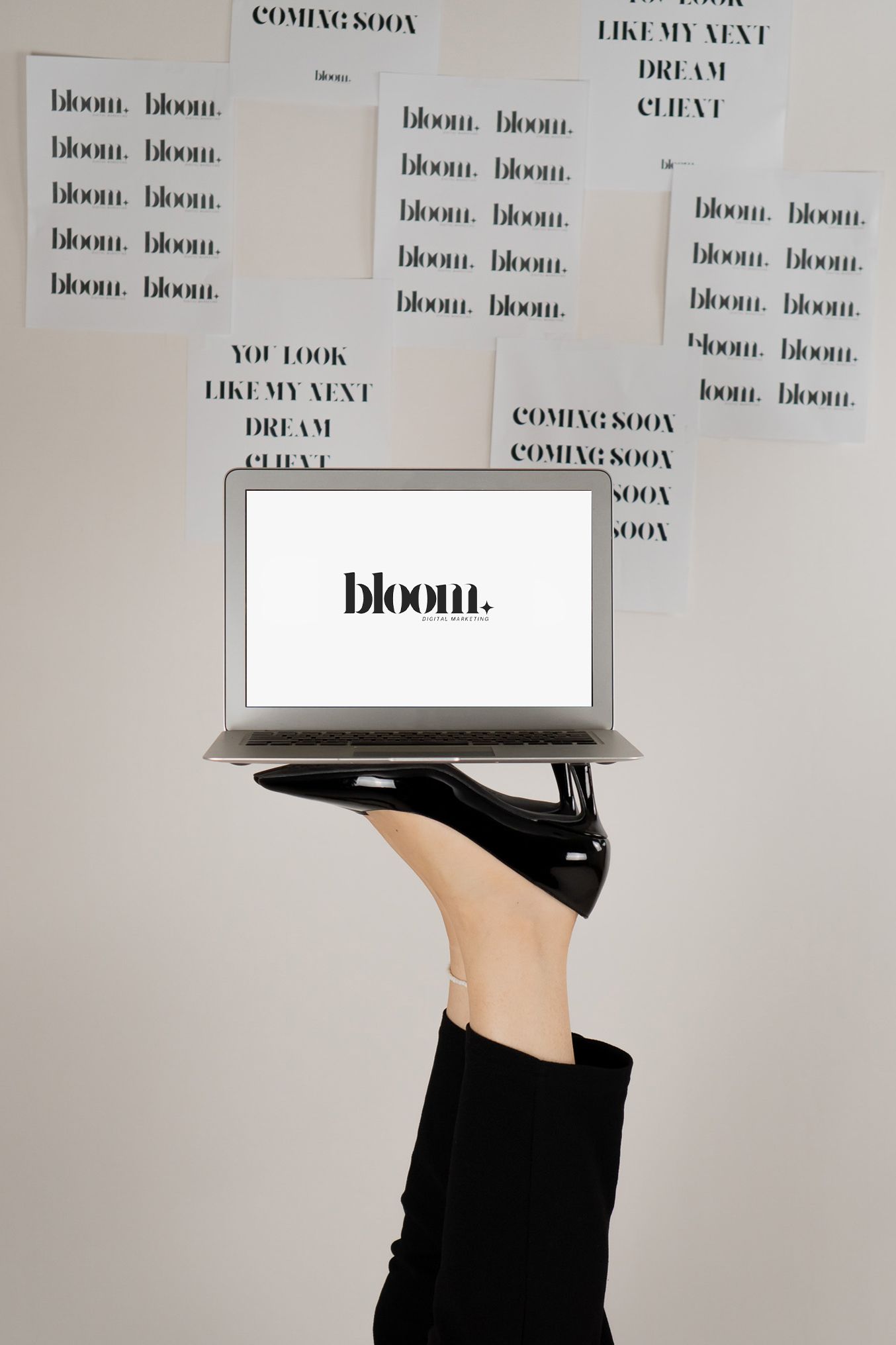 Leg holding laptop displaying bloom logo surrounded by branding papers on white wall.