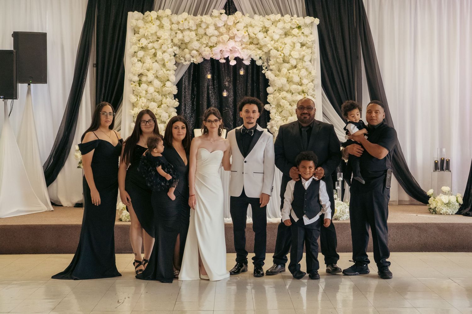 Wedding party posing together in front of a floral arch backdrop at a reception venue.