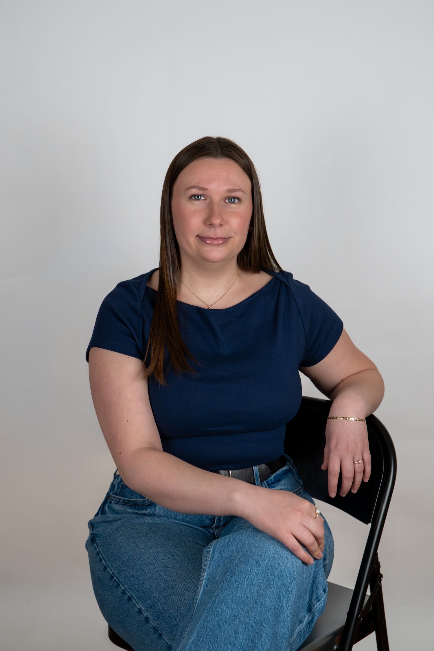 Woman in navy top and jeans sitting on black chair against white background, smiling at camera Headshot photoshoot in New London Connecticut