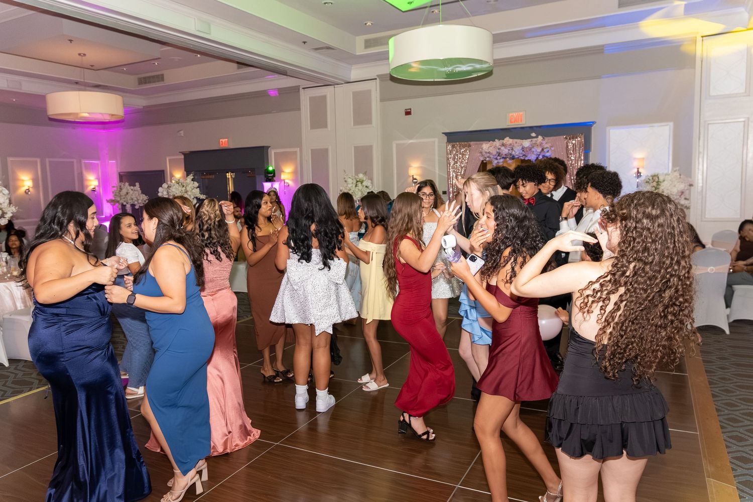 Teens dancing at a formal event in an elegant banquet hall with colorful lighting and floral decorations. Sweet 16!