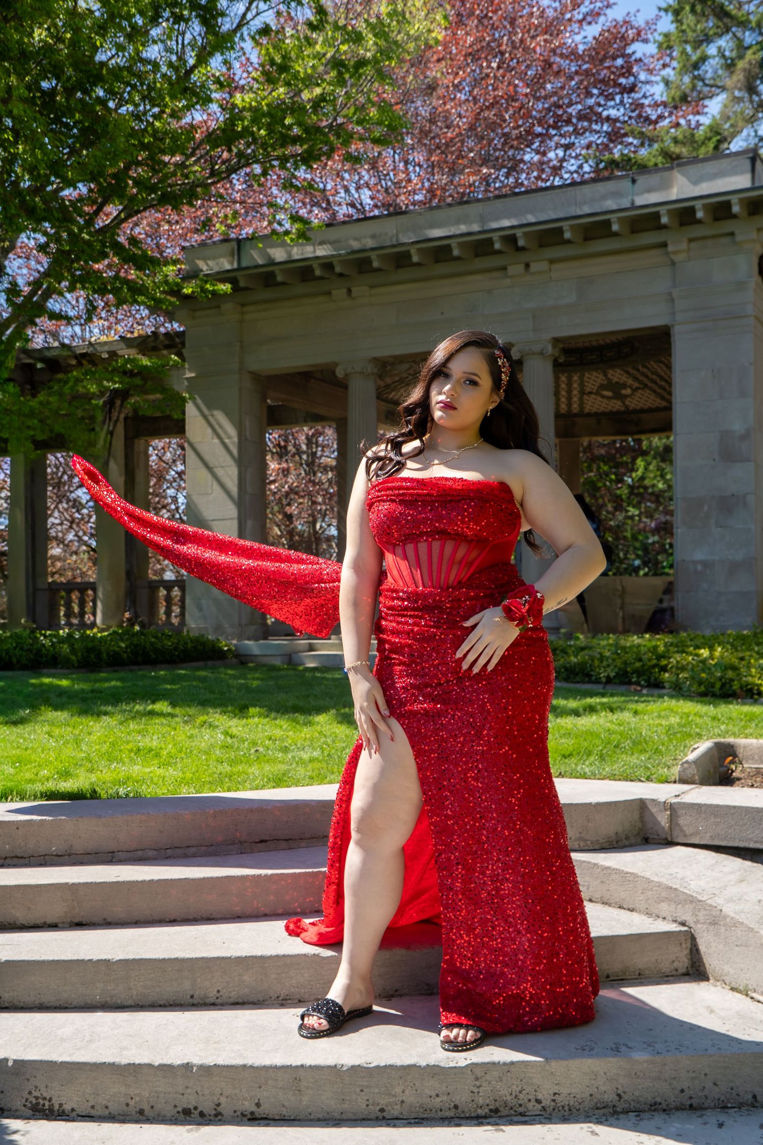 Woman in a stunning red sequin gown with flowing cape poses on stone steps in a garden setting.