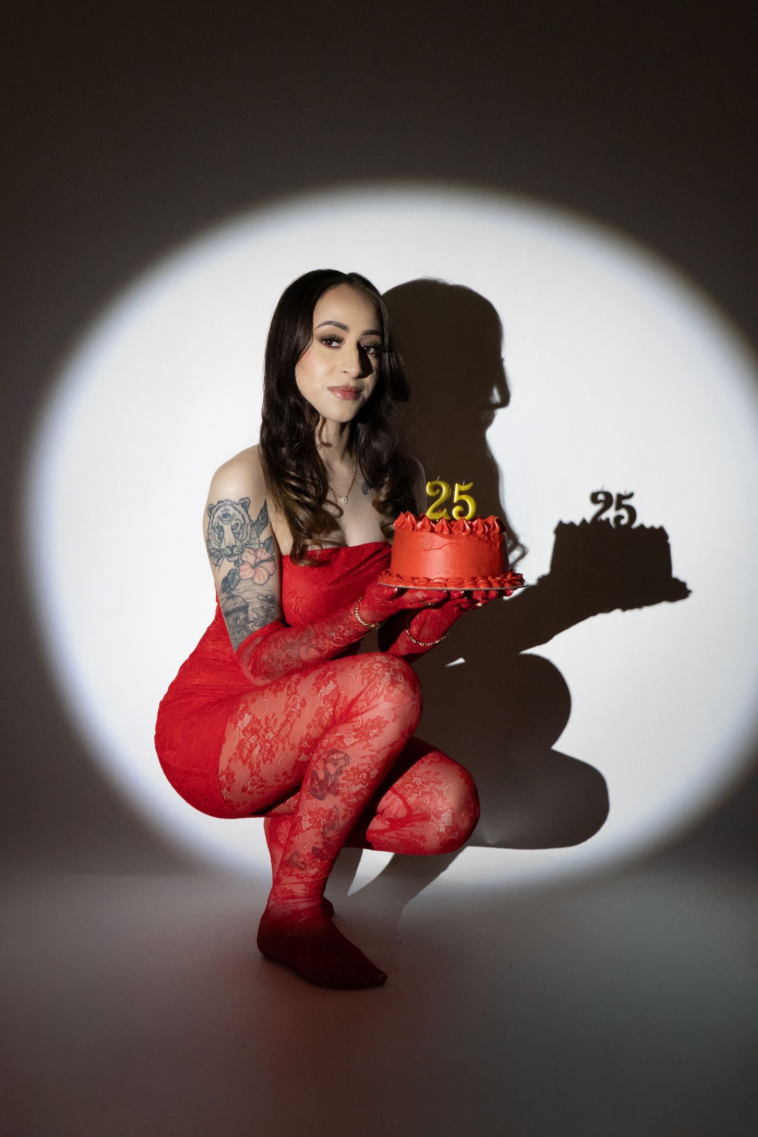 Woman in red lace outfit crouching while holding a red 25th birthday cake.