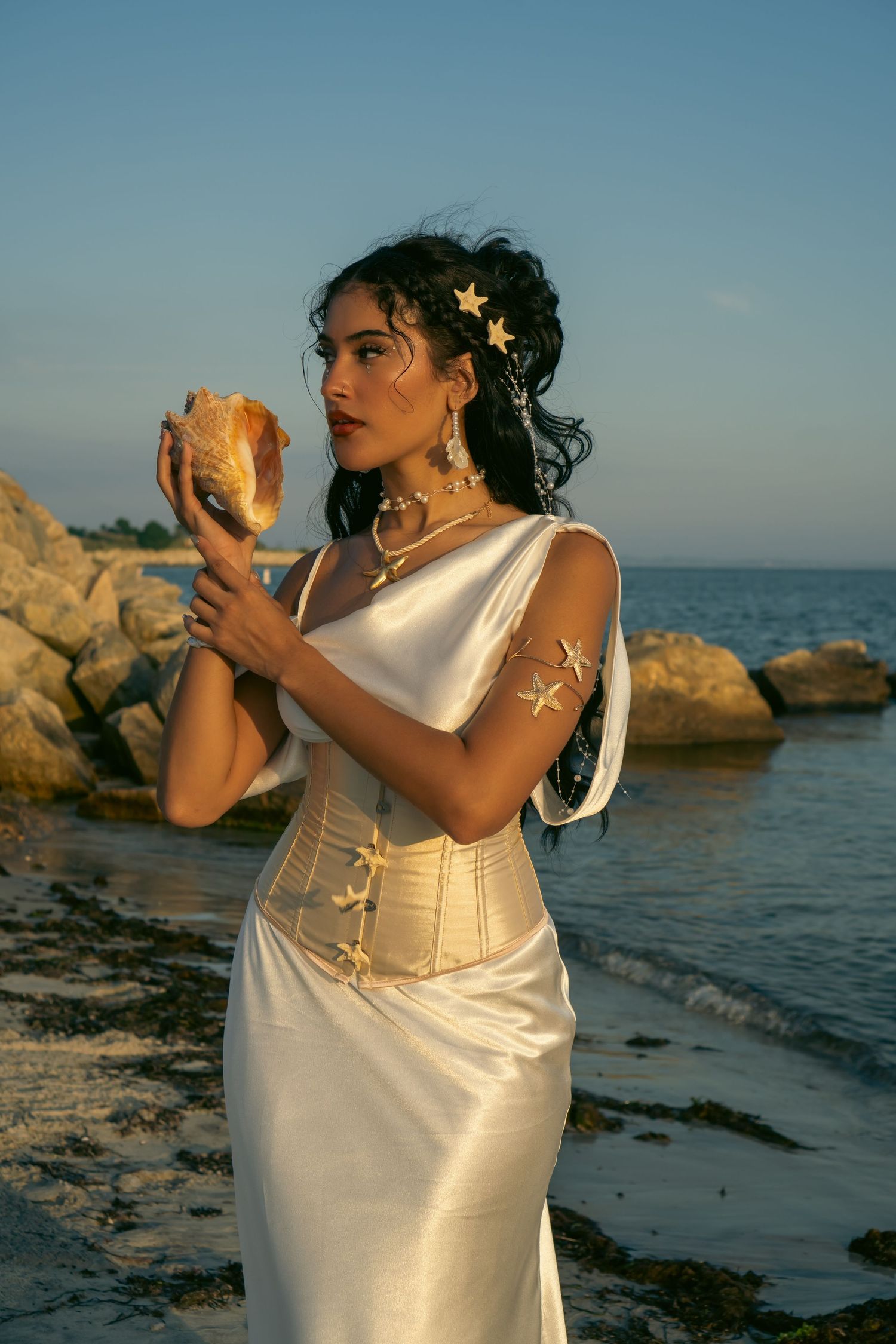 Woman in white satin gown and gold corset holding a seashell on a rocky beach at sunset.