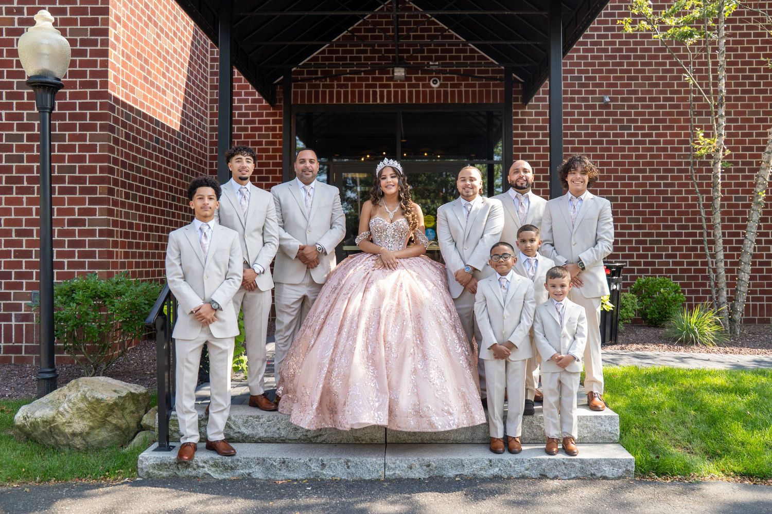 Girl in pink quinceañera gown poses with chambelanes in tan suits on brick building steps. Sweet 16/Quinceañera