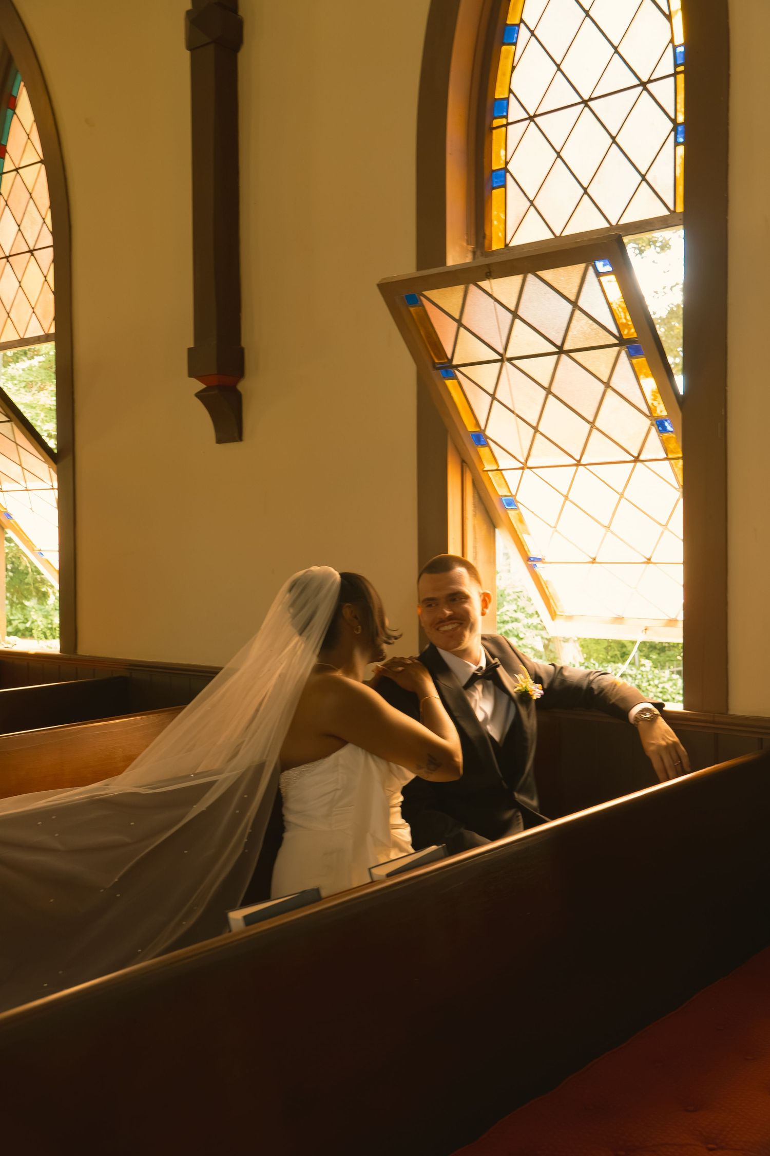 Bride and groom share a romantic moment in a church pew, with beautiful stained glass windows glowing behind them.
