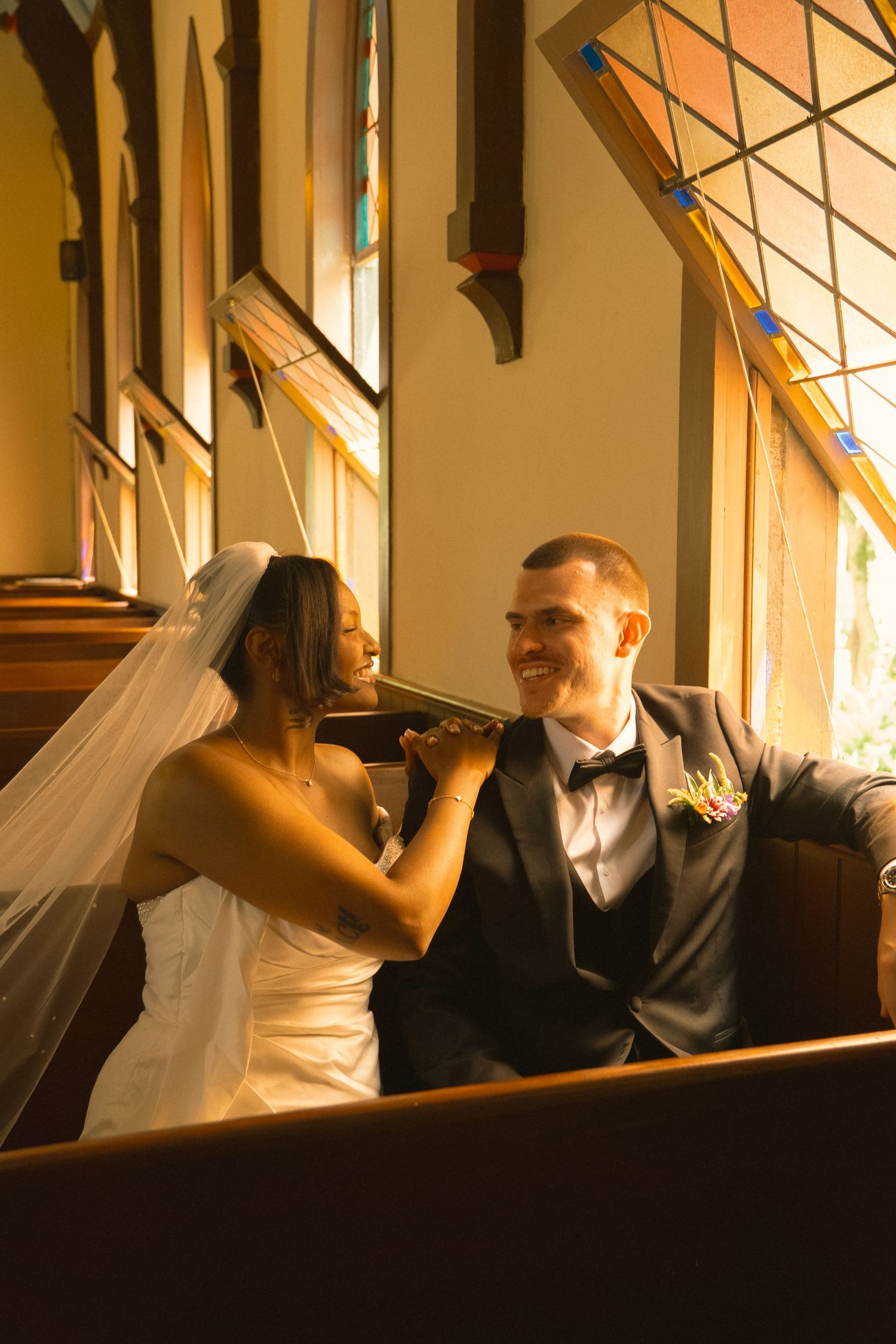 Interracial newlyweds sit together in church pew, bride in white dress and veil, groom in navy suit with boutonniere.
