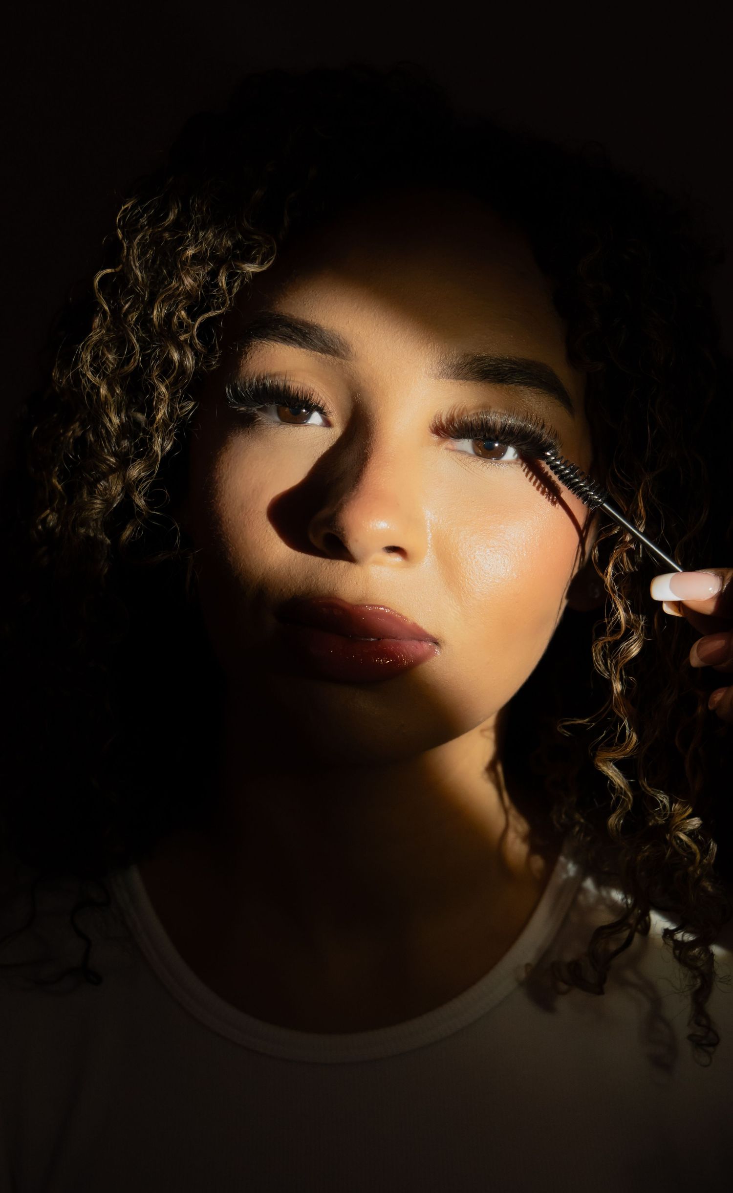 Young woman with curly hair and dramatic lashes applies mascara in moody dramatic lighting.